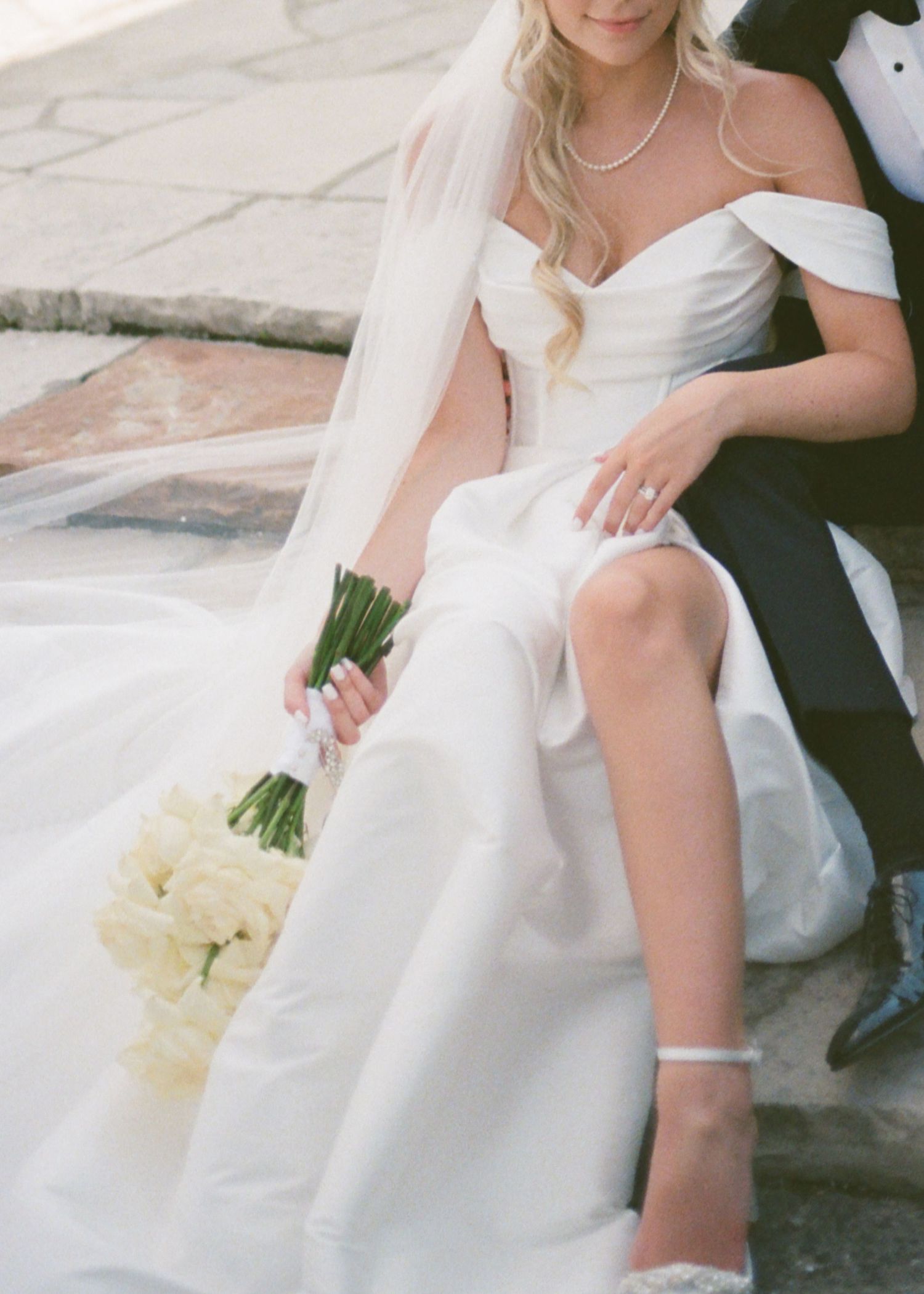 Wedding couple sits together in elegant white gown and formal attire holding bouquet with creamy white roses.