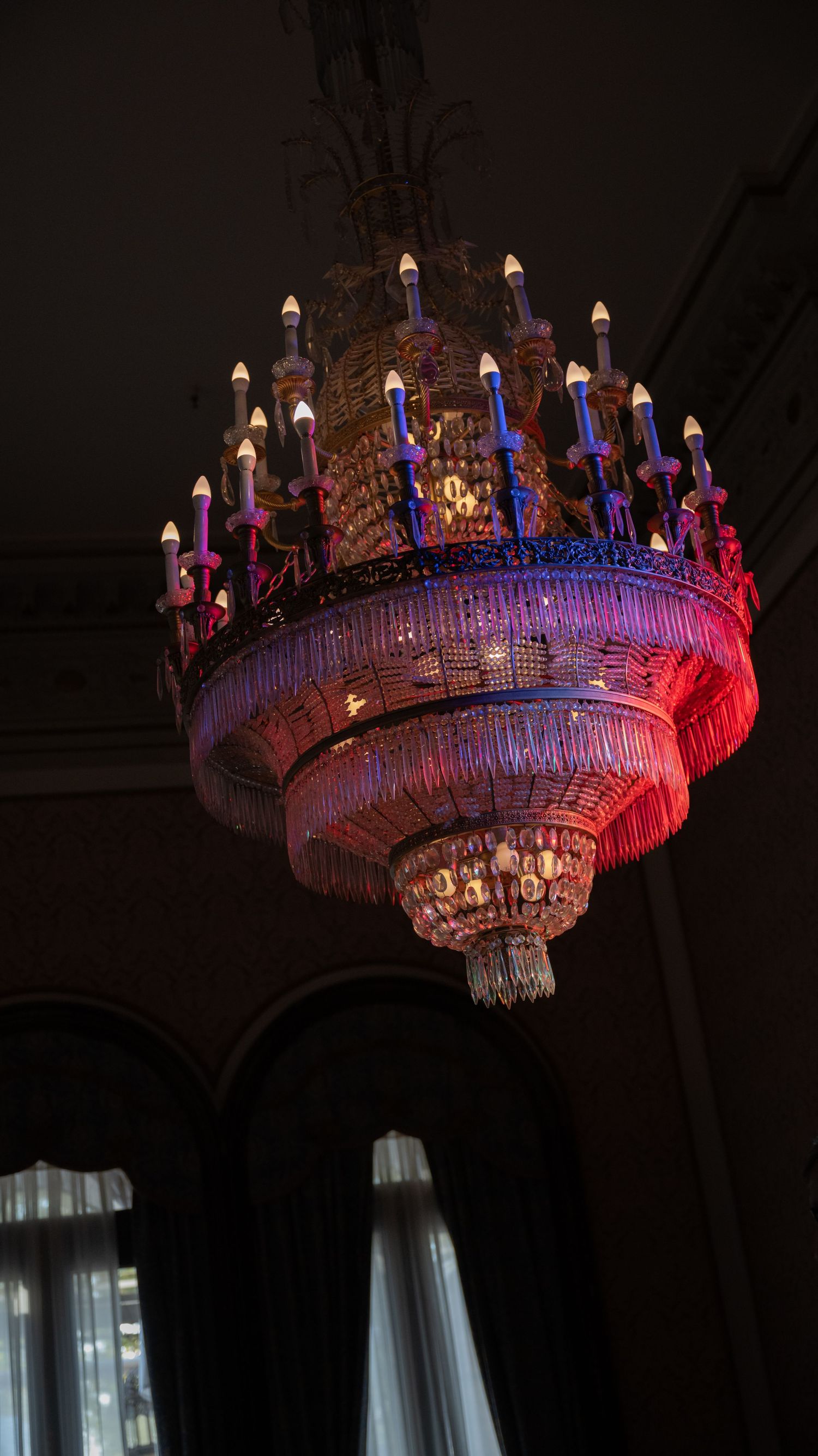 An ornate crystal chandelier illuminated with red and blue lights hangs from a dark ceiling in a dramatic display.