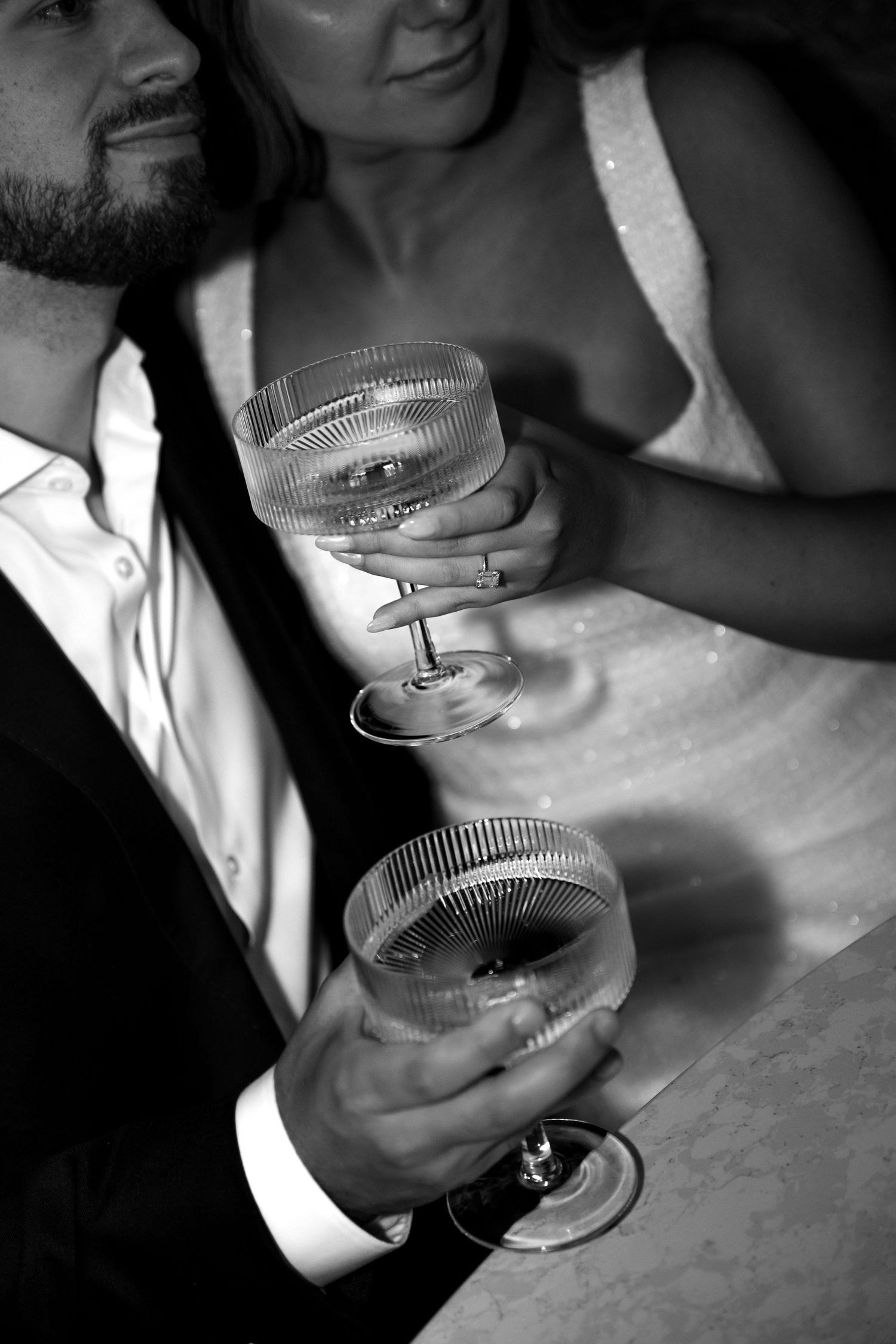 Two hands holding elegant crystal cocktail glasses in a black and white intimate setting.