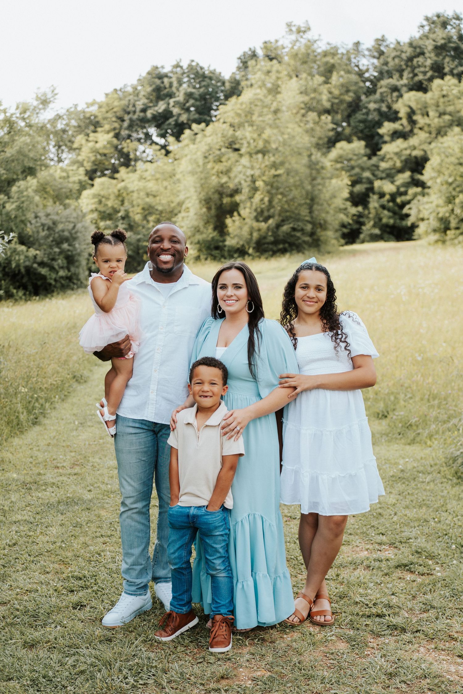 A family poses together in a grassy field on a sunny day wearing coordinated light blue and white outfits.