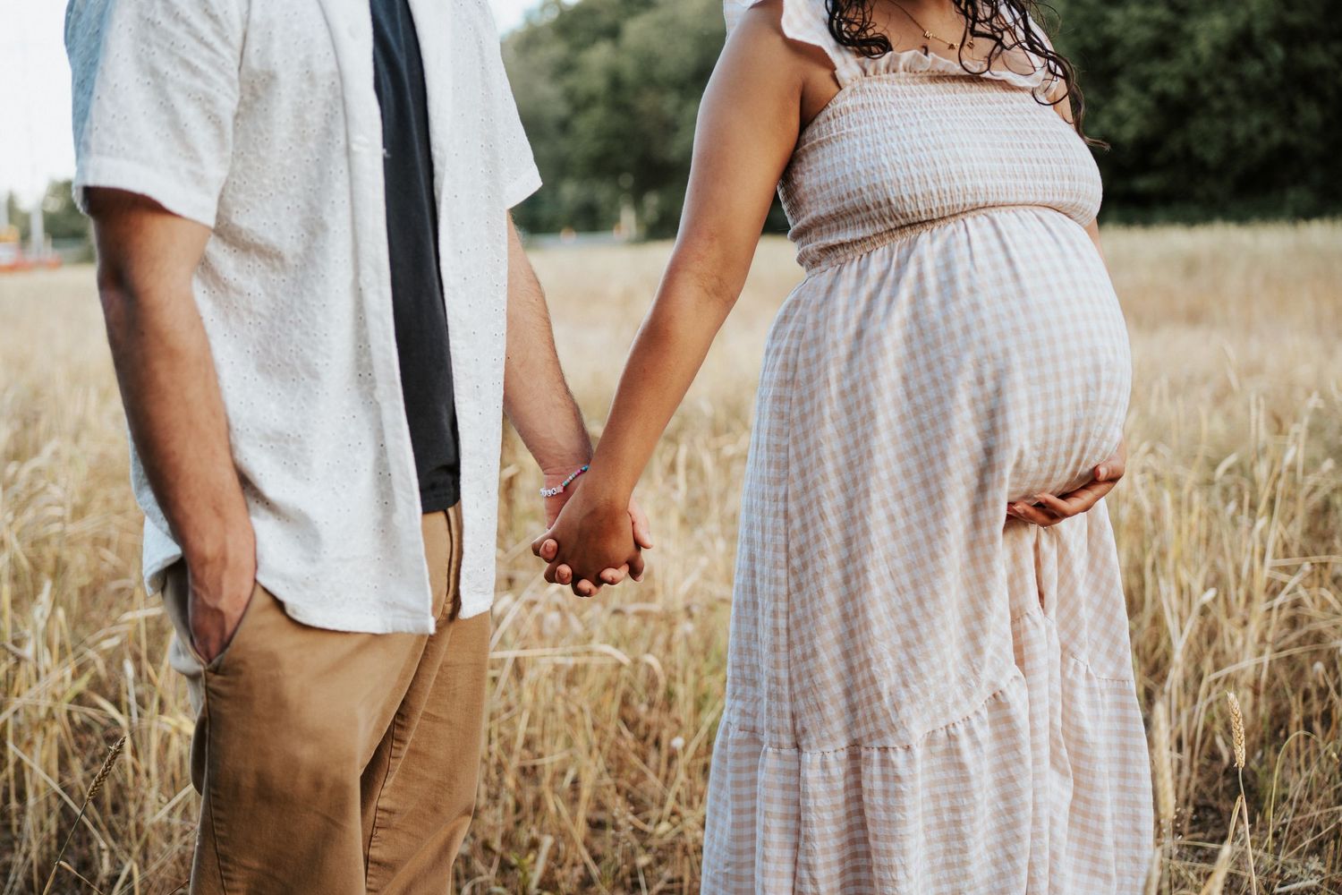 Couple holding hands in wheat field with person wearing white maternity dress and partner in casual attire.