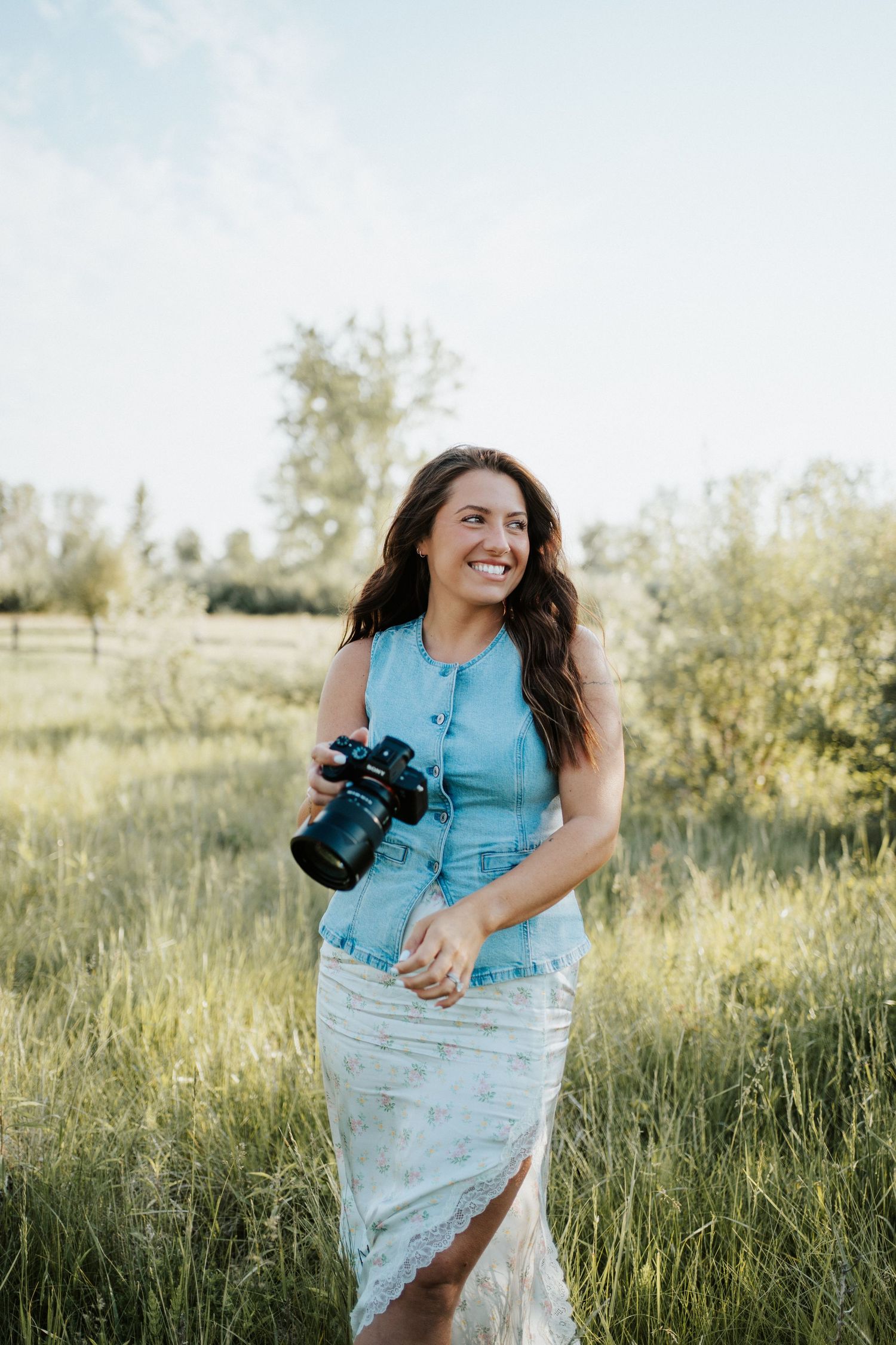 Smiling woman holding a DSLR camera in a green field wearing a blue denim top and floral skirt on a sunny day.