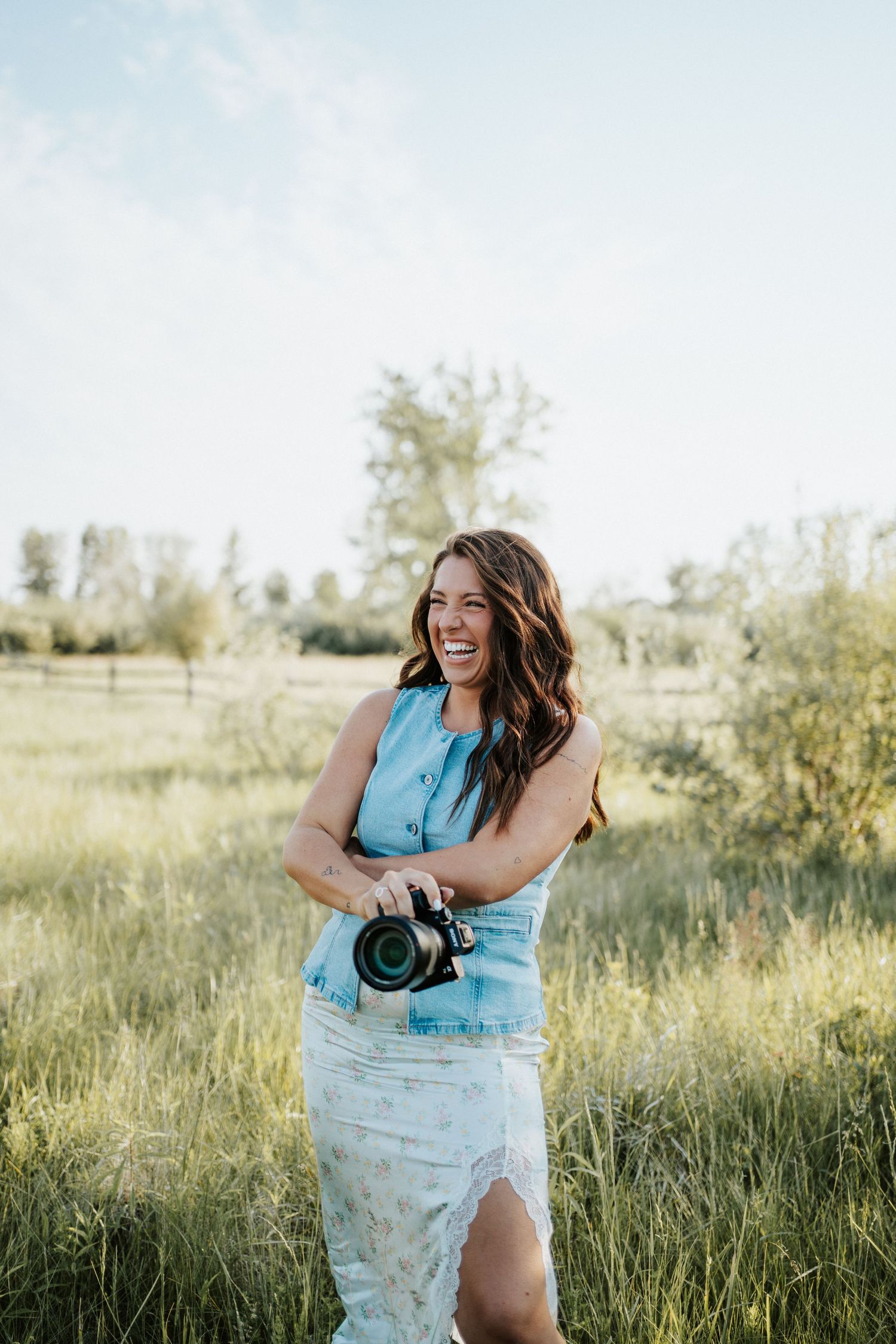 A smiling woman holding a DSLR camera stands in a lush green field wearing a denim top and floral skirt.