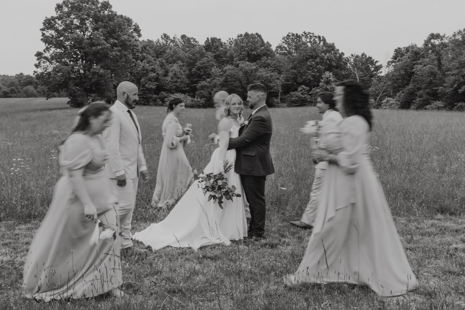 A black and white wedding ceremony taking place in a scenic field with bridesmaids in flowing dresses.