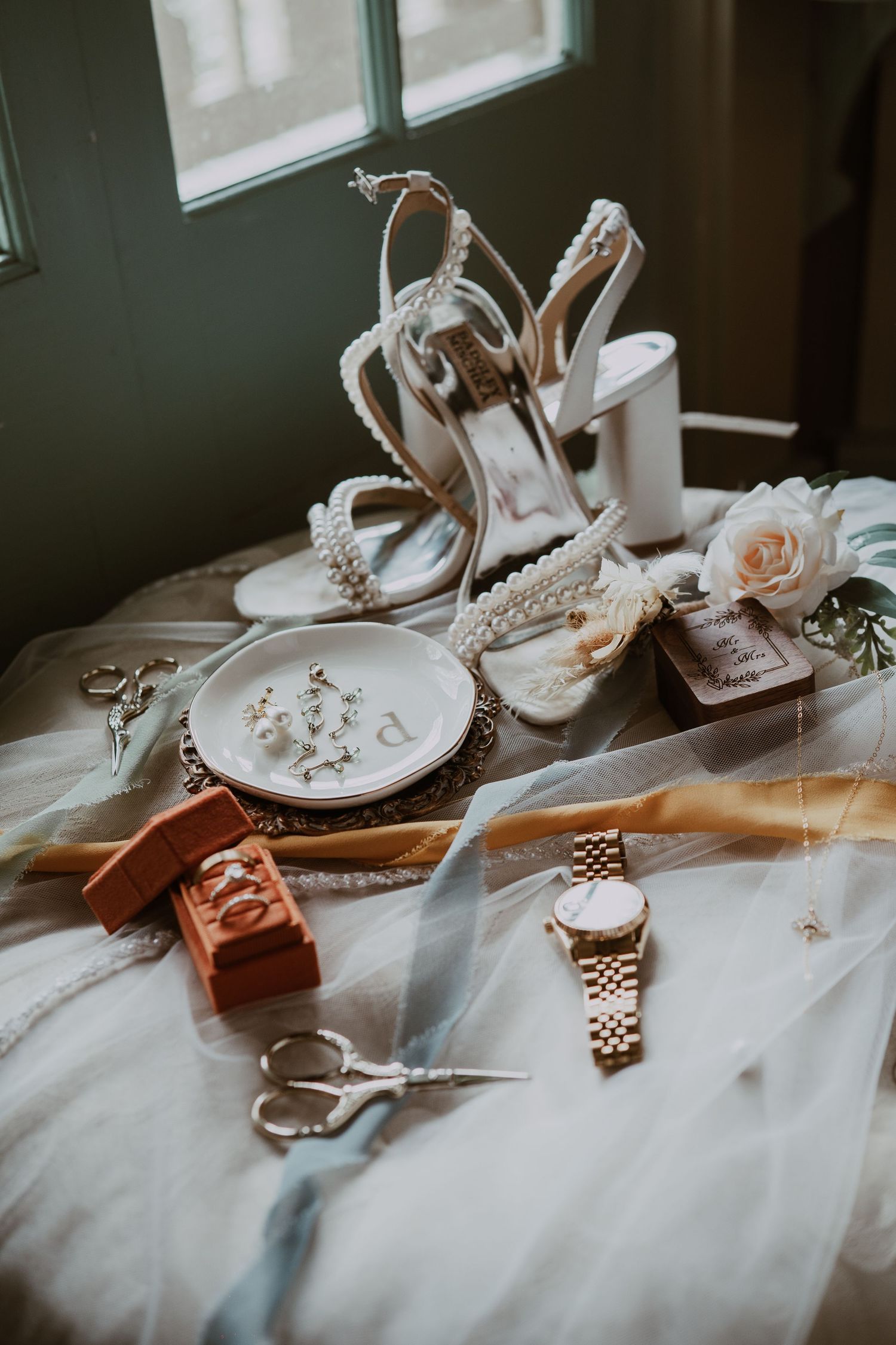 Wedding accessories including white heels, jewelry, watches and roses laid out on a white surface near a window.