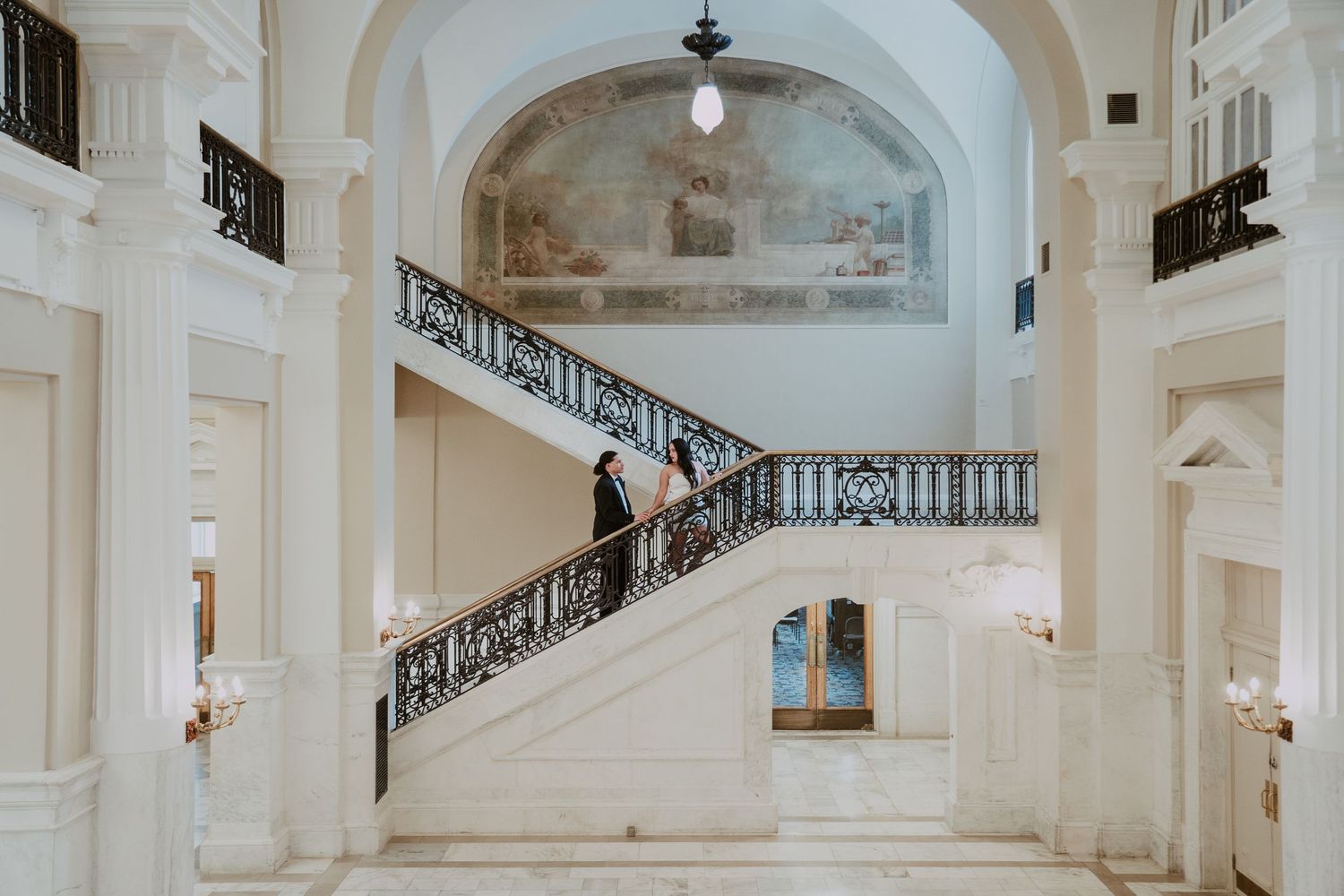 Elegant white marble staircase with ornate black railings in a grand historic building interior with arched ceiling.
