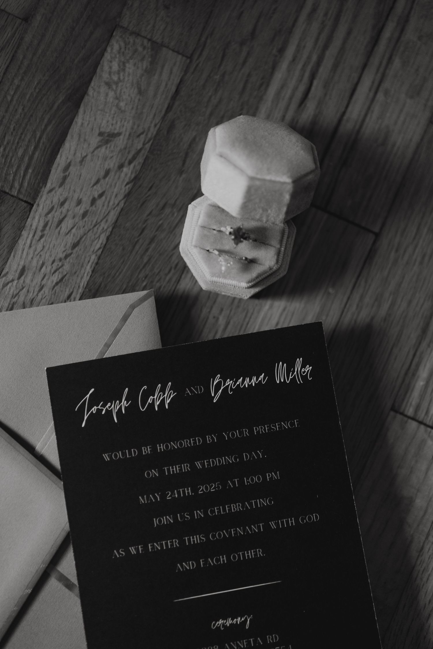 A black and white wedding invitation sits beside a white geometric ring box on a wooden surface.