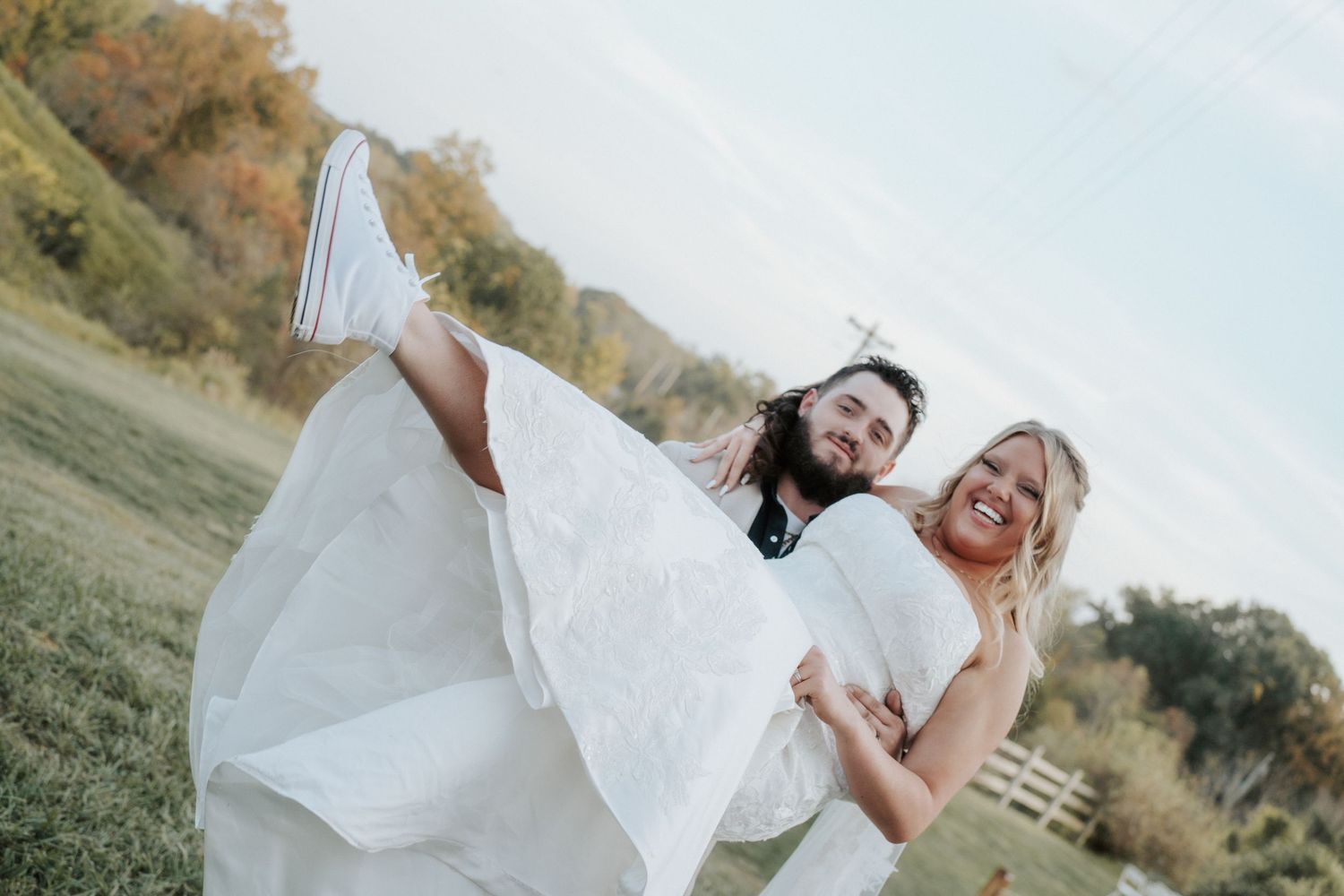 Joyful groom carrying bride in white wedding dress outdoors on green field with autumn trees in background.