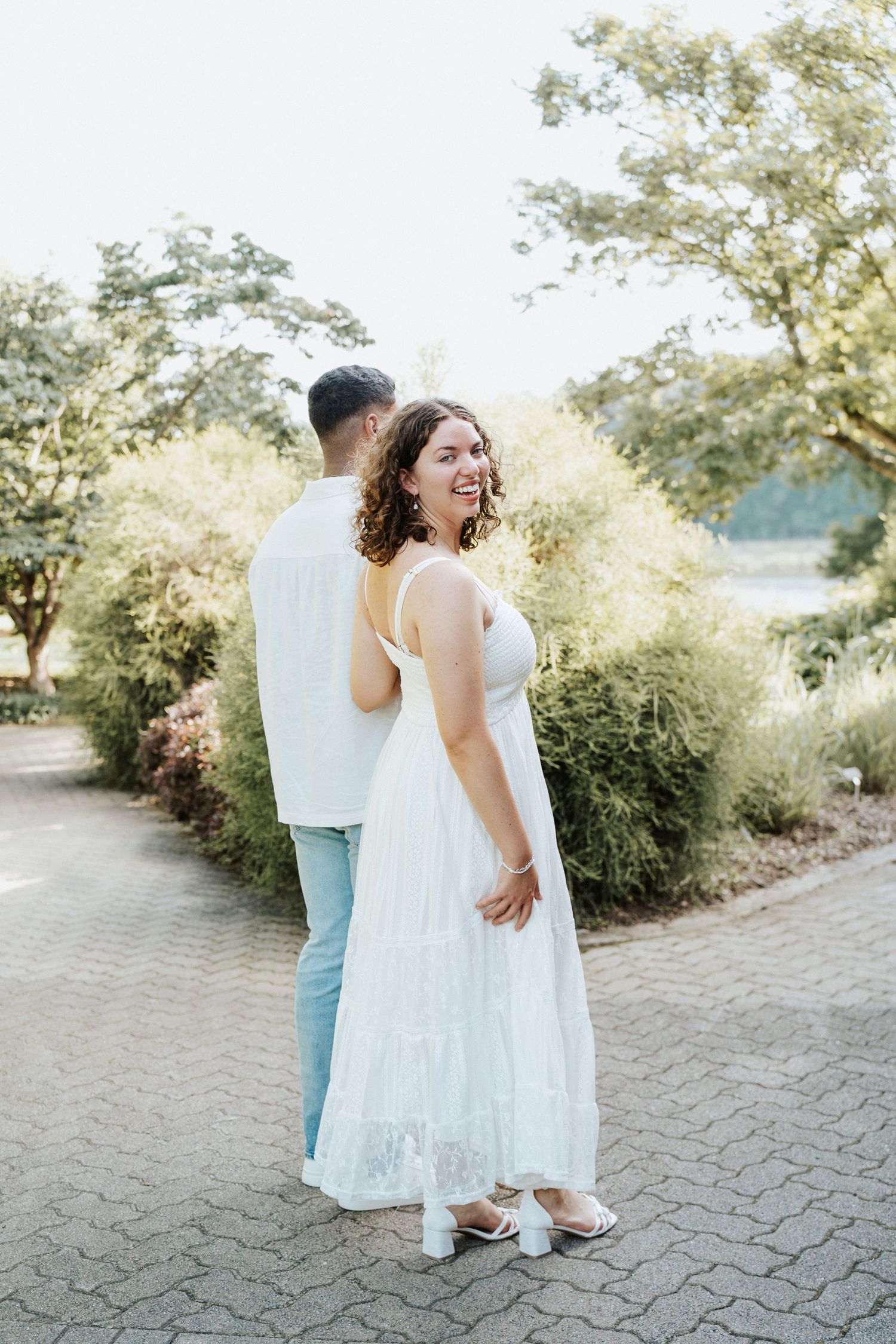 Two people in light colored outfits share a joyful moment together while dancing on a stone pathway surrounded by greenery.
