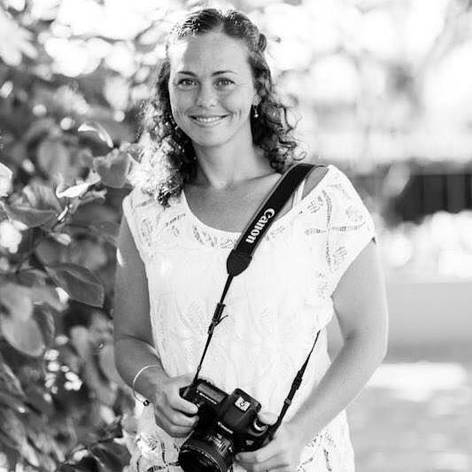 A smiling woman in a white lace dress holds a Canon camera outdoors in a black and white portrait photo.