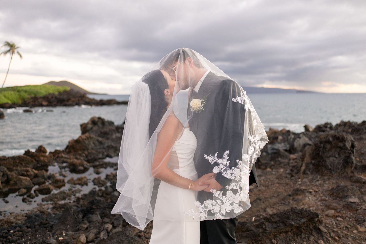 Bride and groom sharing a romantic kiss under a lace veil on rocky Hawaiian coastline at sunset.