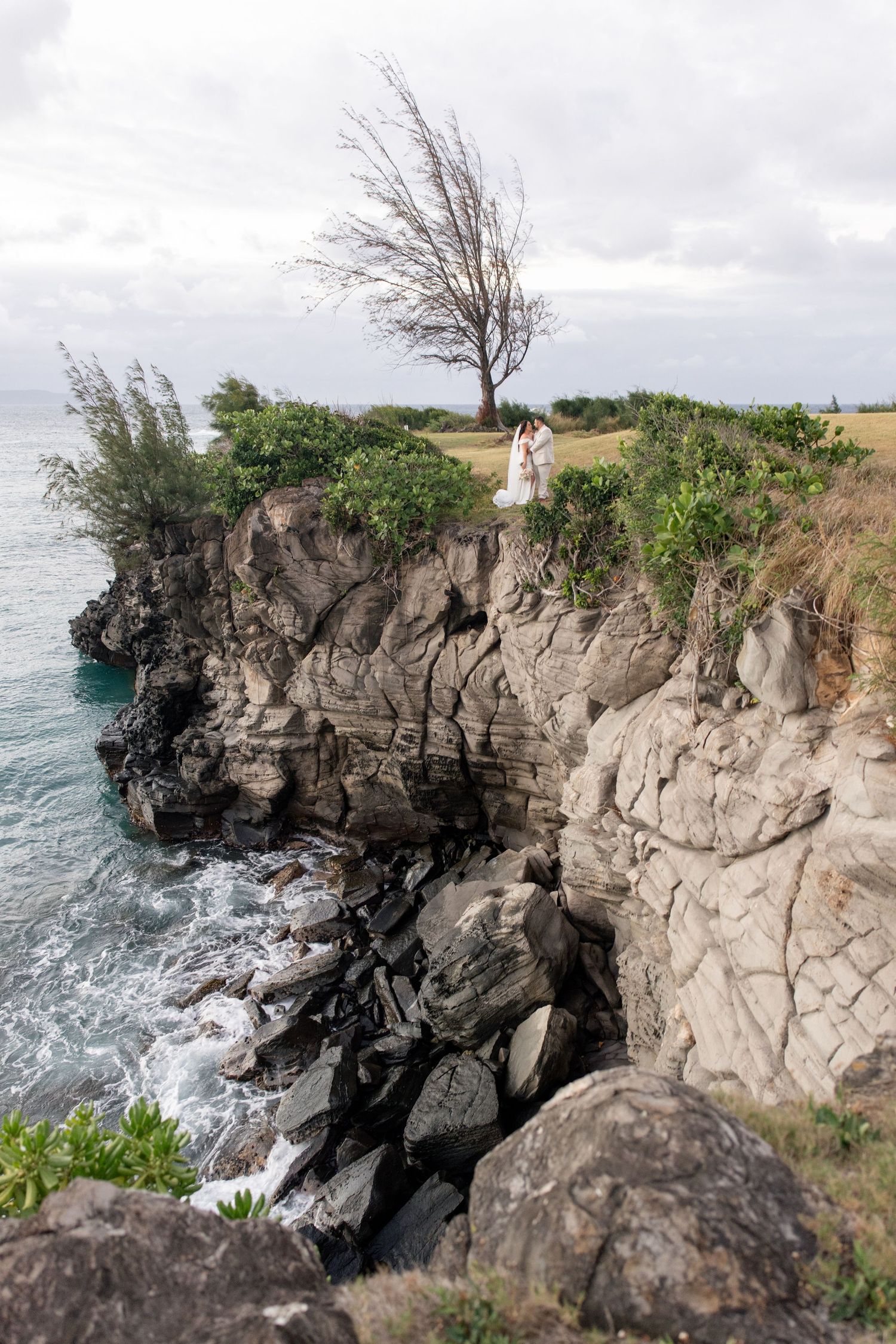 Couple embracing on rocky coastal cliffs with turquoise ocean waves, lush greenery, and a windswept tree under cloudy skies.
