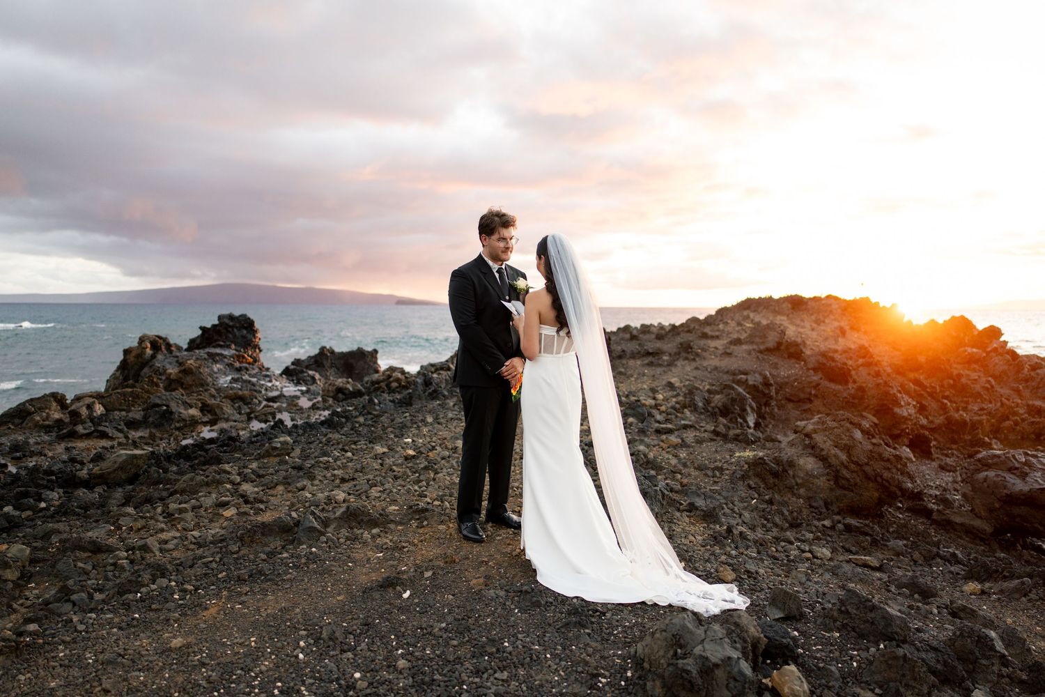 Bride and groom embrace on volcanic rocks at sunset with ocean views in Maui, Hawaii wedding photo.