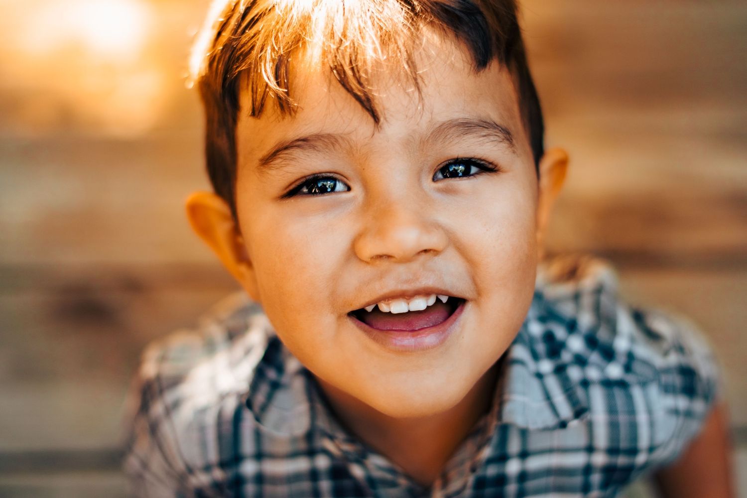 A 2 yo boy with tan skin and brown hair smiling up at the camera. Sunlight is shining through his hair and sparkling in 