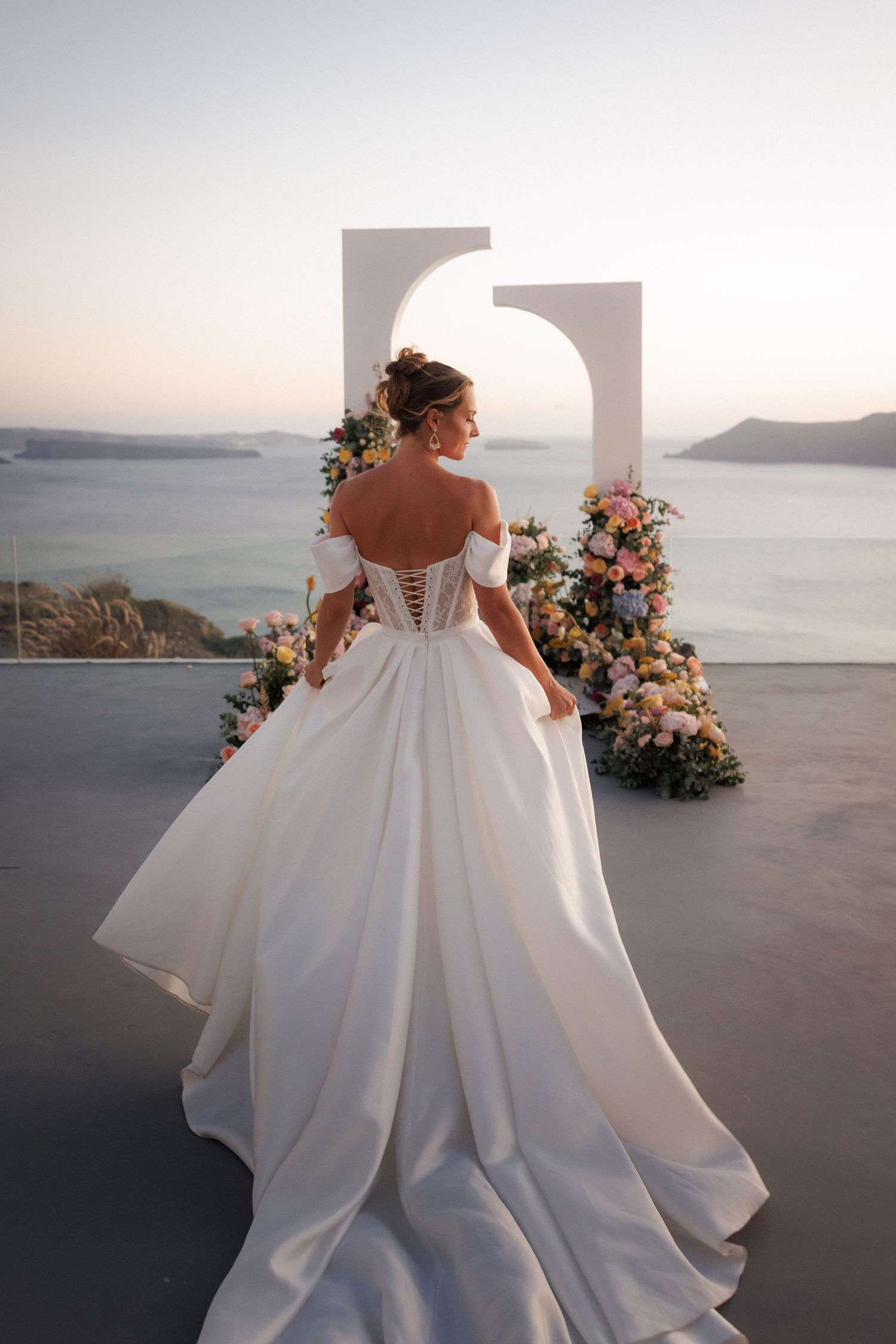 A bride in an elegant white off-shoulder gown stands beside floral arrangements on a terrace overlooking the Mediterranean sea.