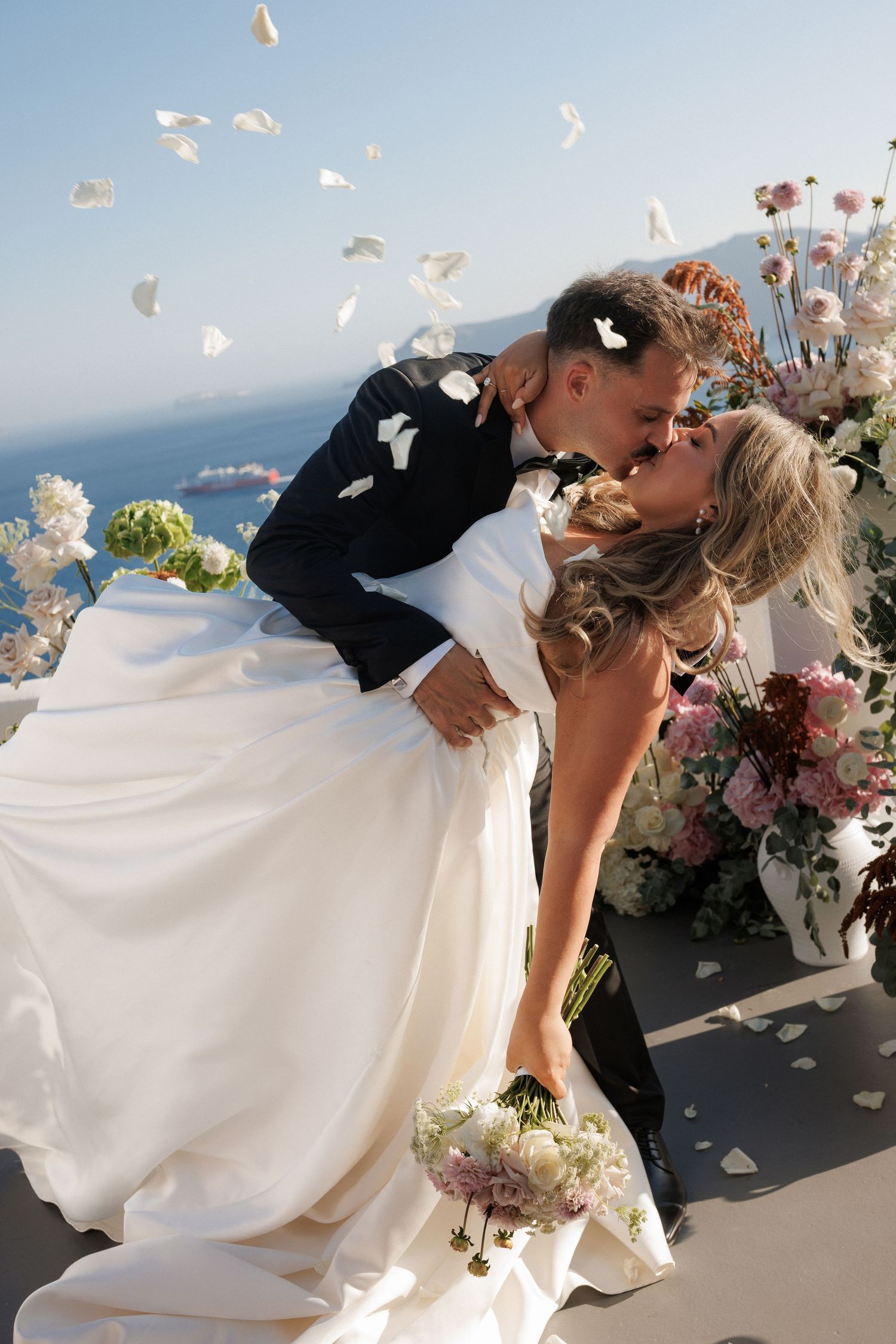 Romantic wedding scene in Santorini with flower petals floating in the air against blue Mediterranean backdrop.