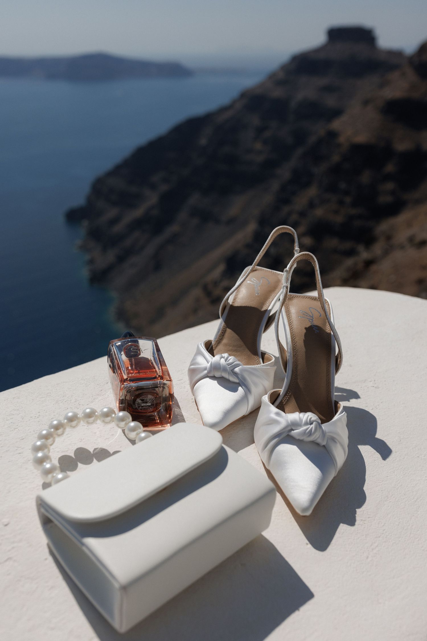White bridal accessories including shoes and clutch displayed on white ledge overlooking Santorini's dramatic caldera views.