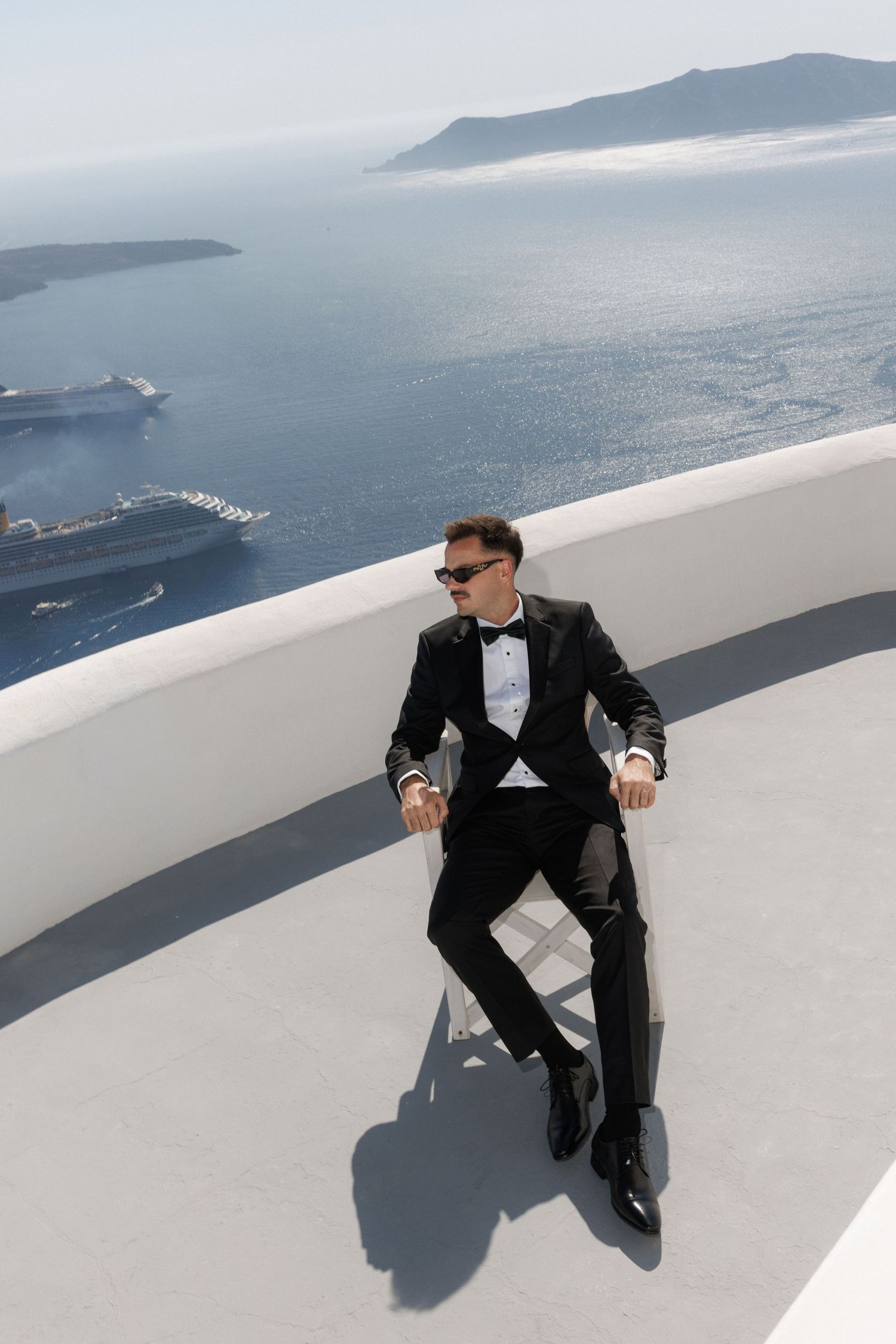 Person in formal black tuxedo sits on white wall overlooking scenic Mediterranean coastline and blue ocean in Santorini.