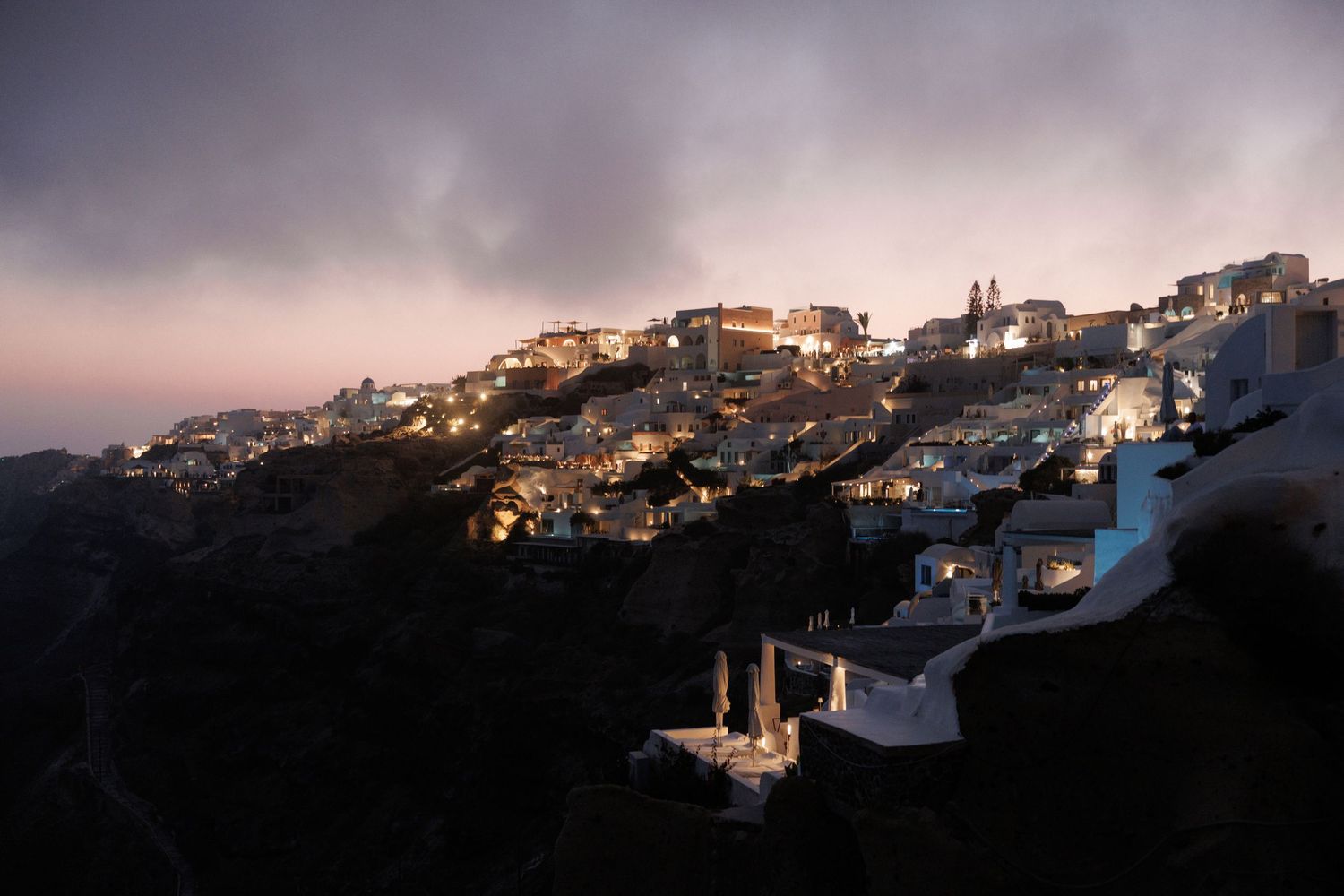 Scenic nighttime view of the illuminated hillside town of Santorini Greece with white buildings glowing against dark sky.