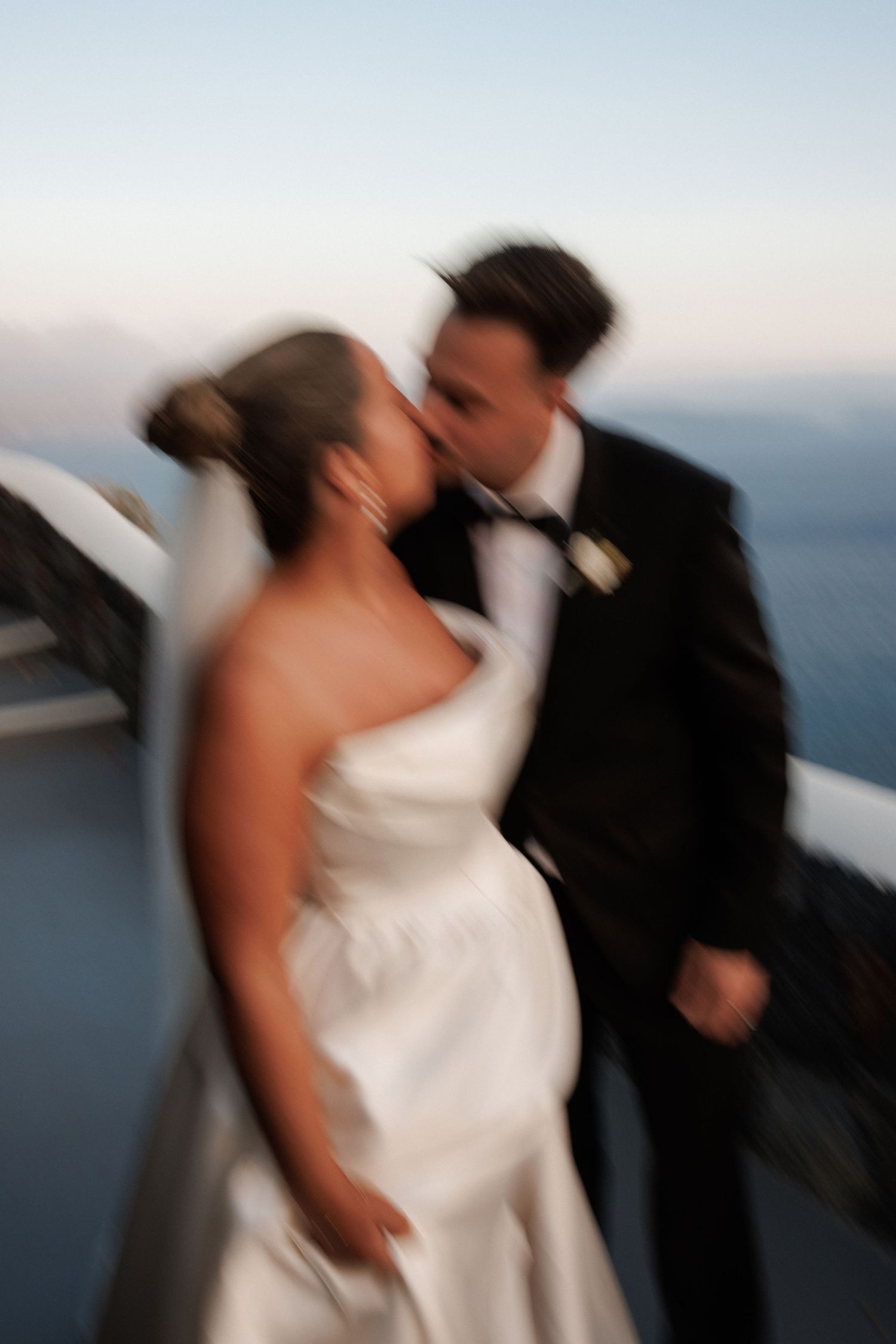 A blurred motion wedding photograph capturing a romantic kiss against a soft blue horizon backdrop.