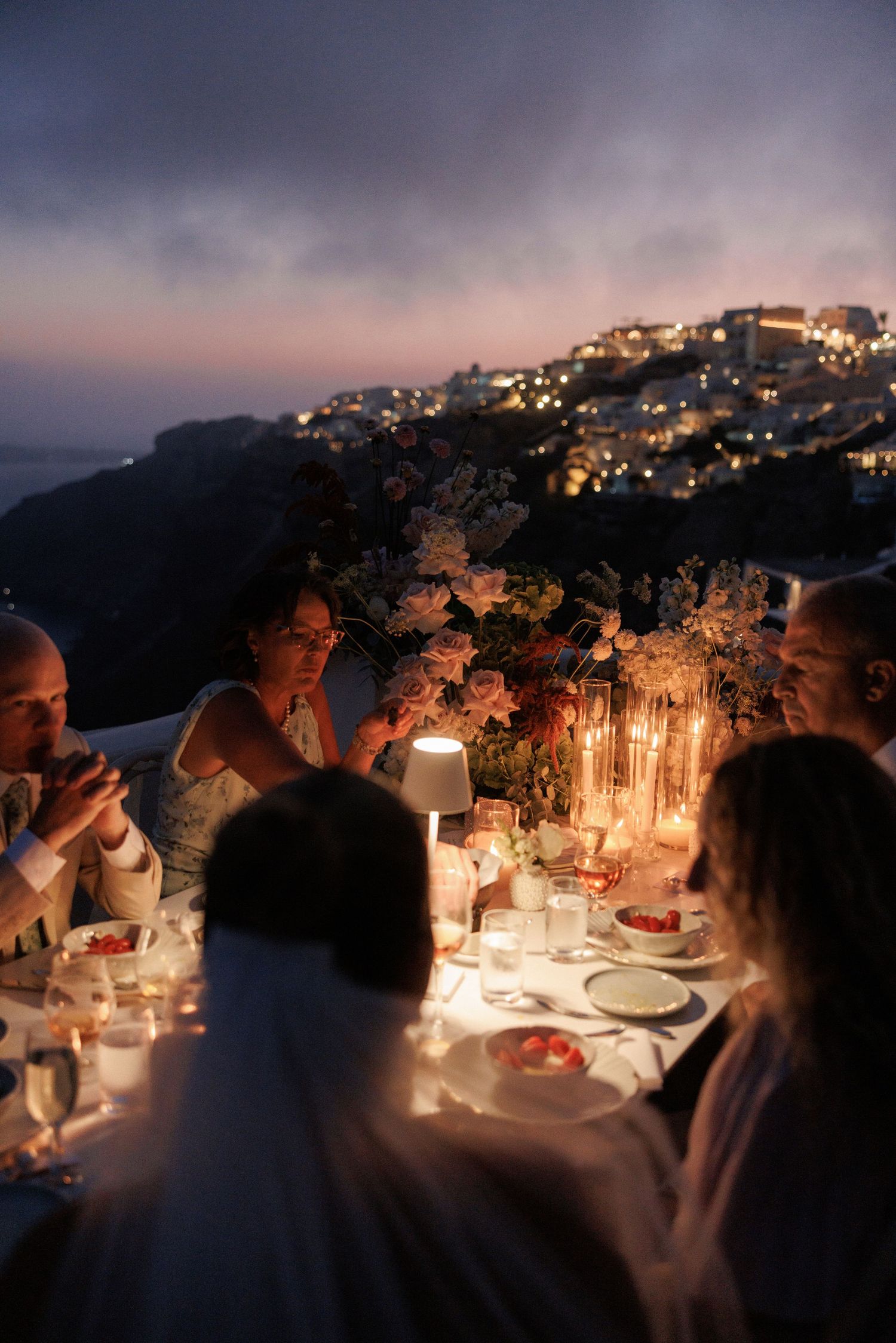 Candlelit outdoor dining tables at dusk create a romantic ambiance with city lights glowing in the distant hillside.