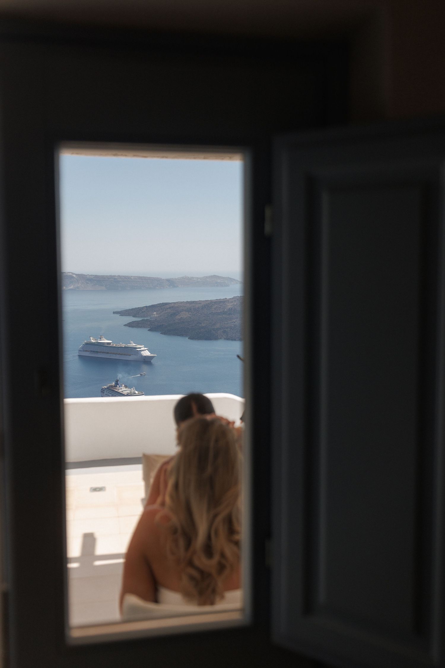 View through open window in Santorini shows cruise ships anchored in blue Mediterranean waters below white buildings.