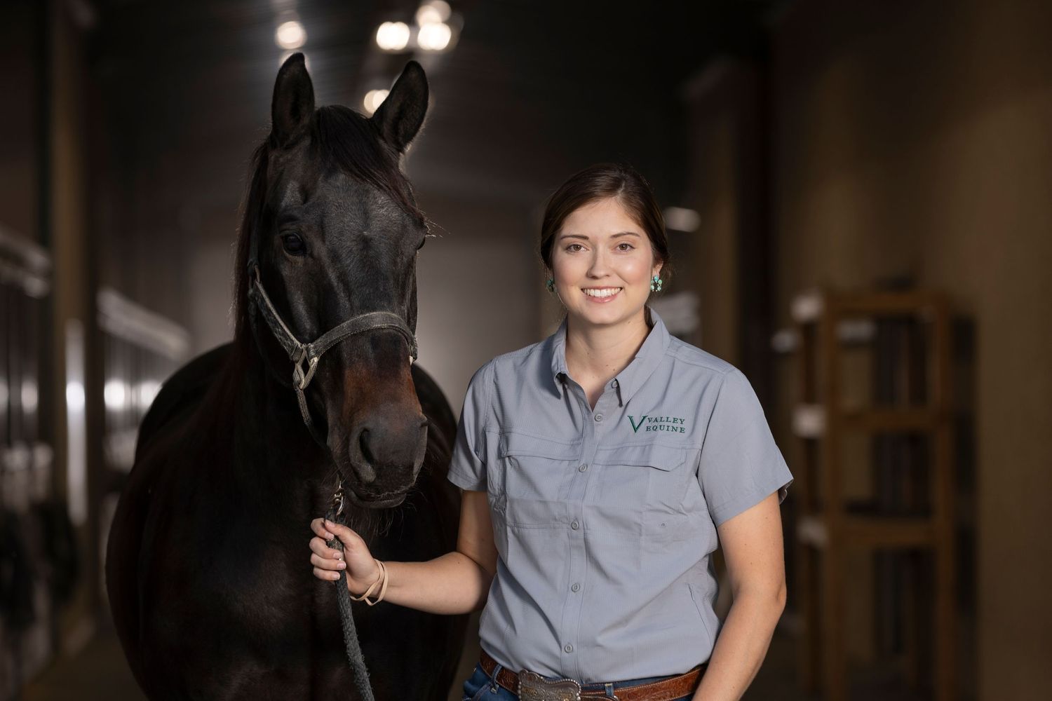 A person in a gray polo shirt stands with a majestic black horse in a dimly lit stable hallway.