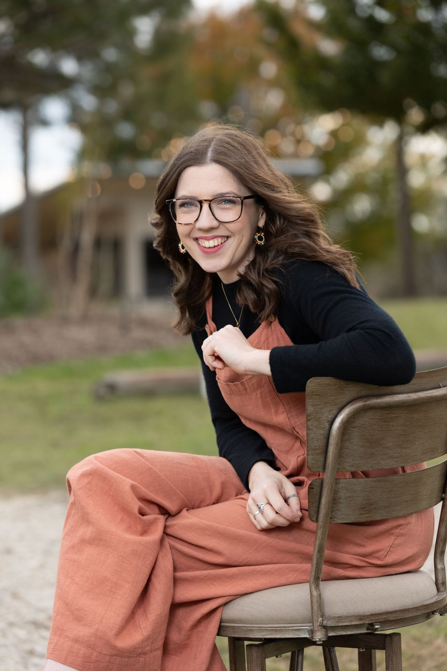 Person smiling while sitting in rustic chair outdoors wearing orange jumpsuit and black sweater.