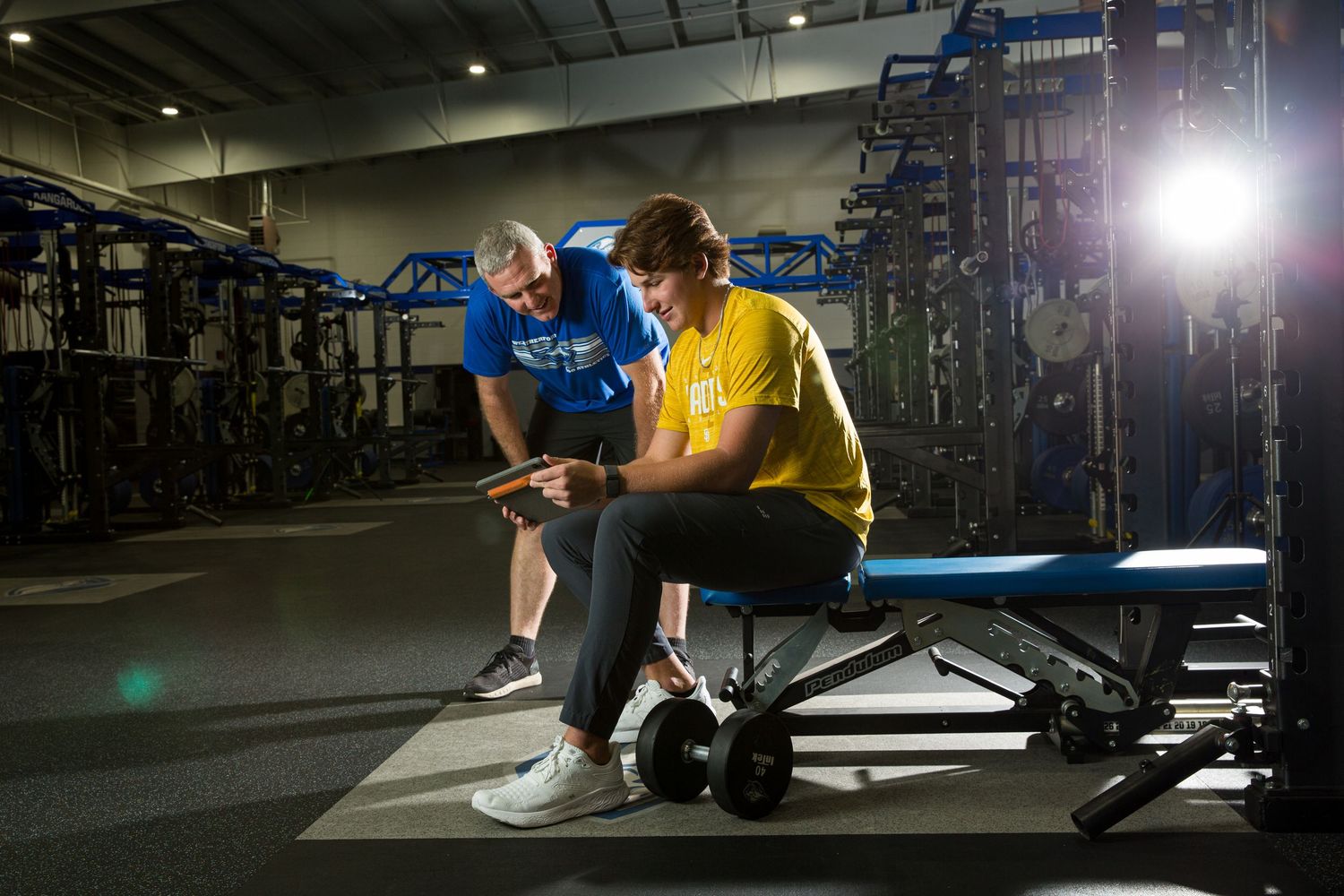 Personal trainer and client reviewing workout progress on a digital device in a dimly lit gym setting.