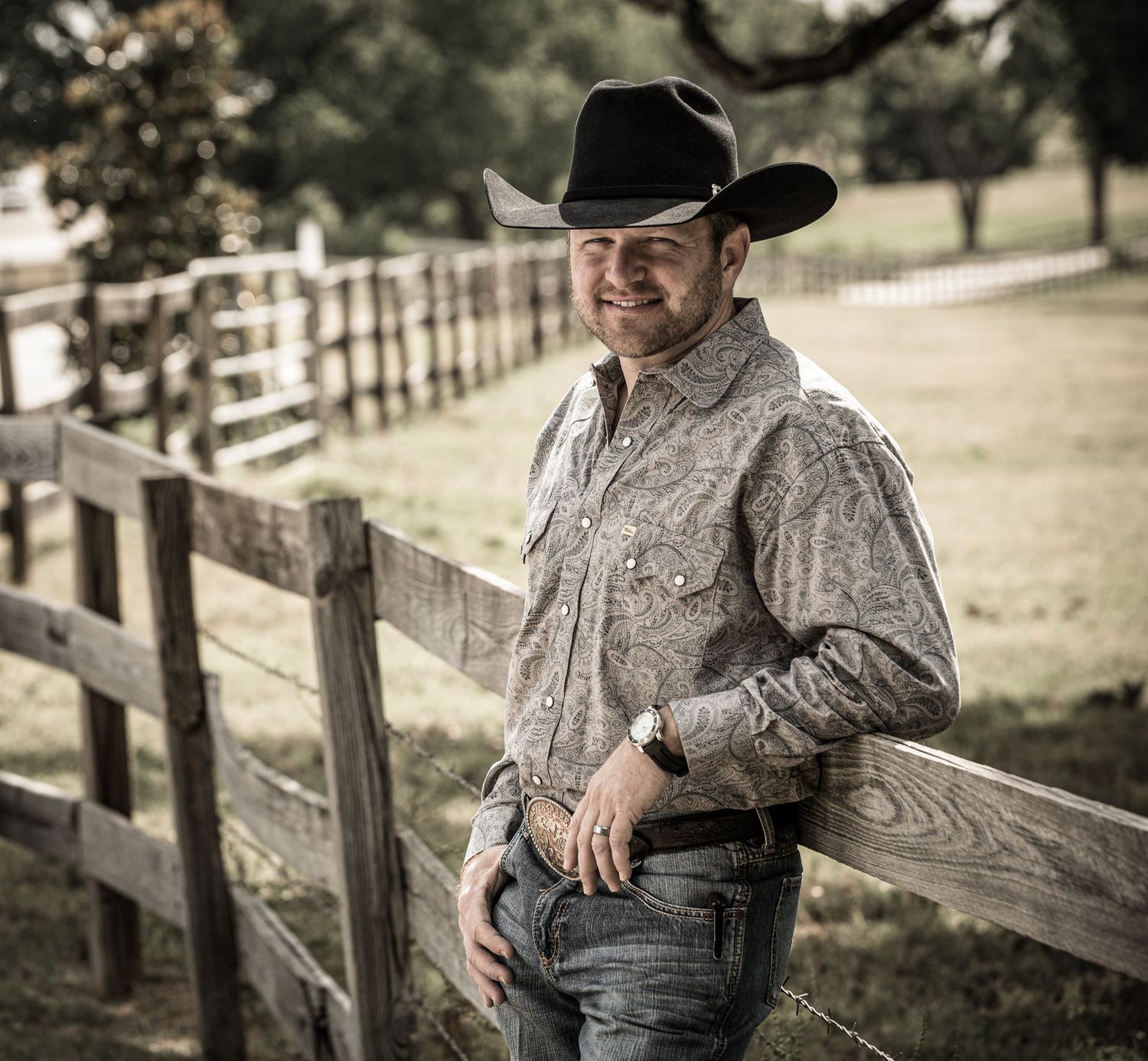 A cowboy wearing a black hat and paisley shirt leans against a wooden fence on a ranch.
