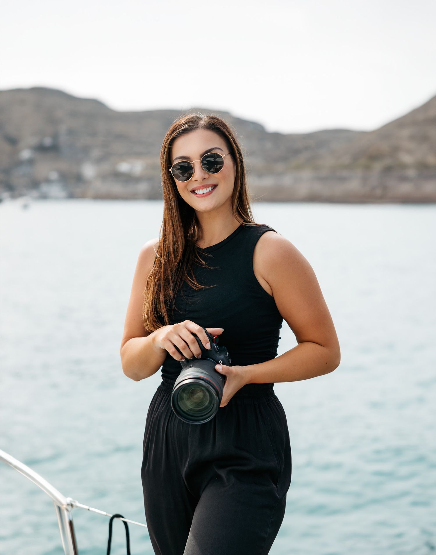 A photographer in black attire poses with camera on sailboat with coastal scenery.