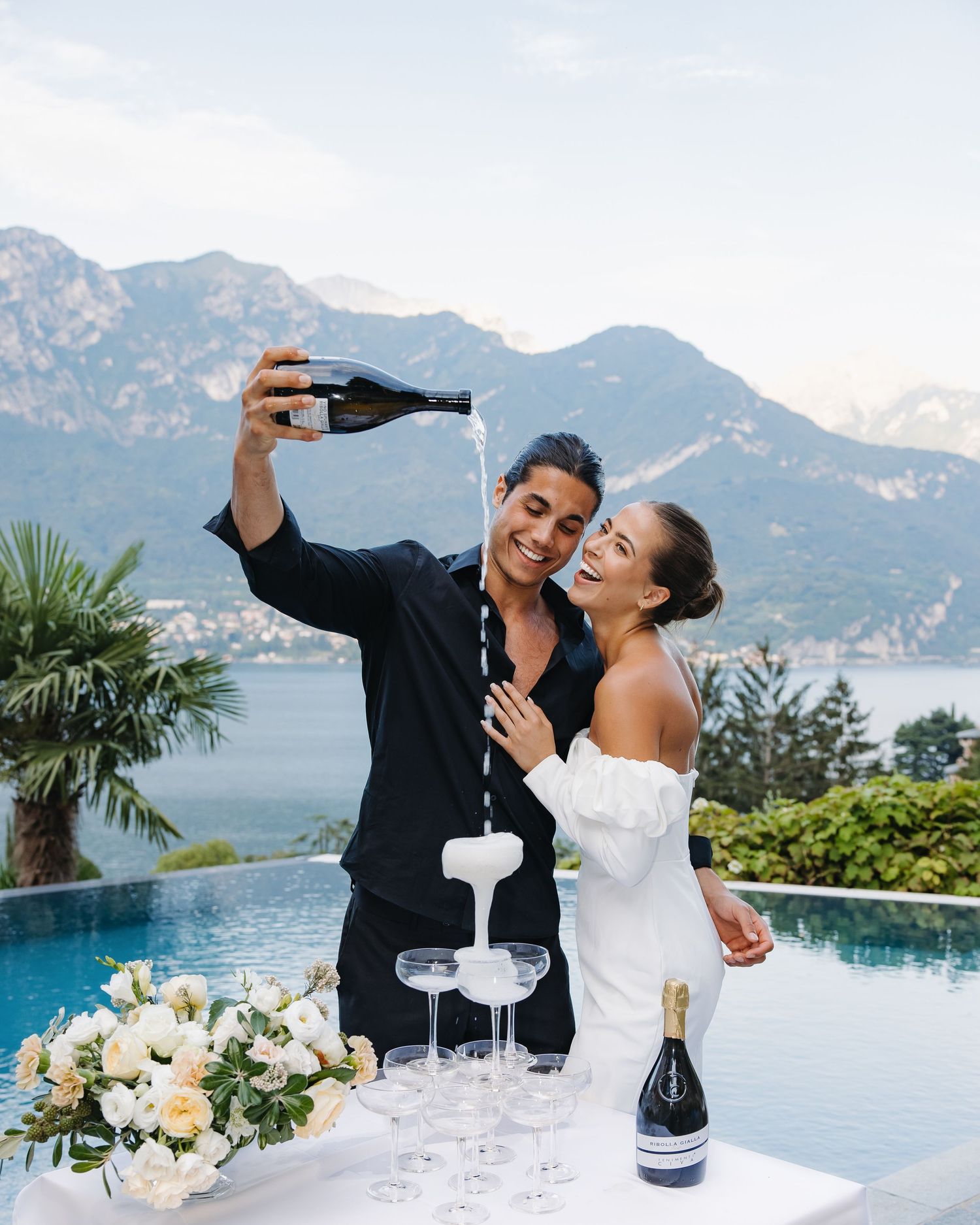 Couple pouring champagne into tower of glasses by infinity pool with mountain and lake como in background.