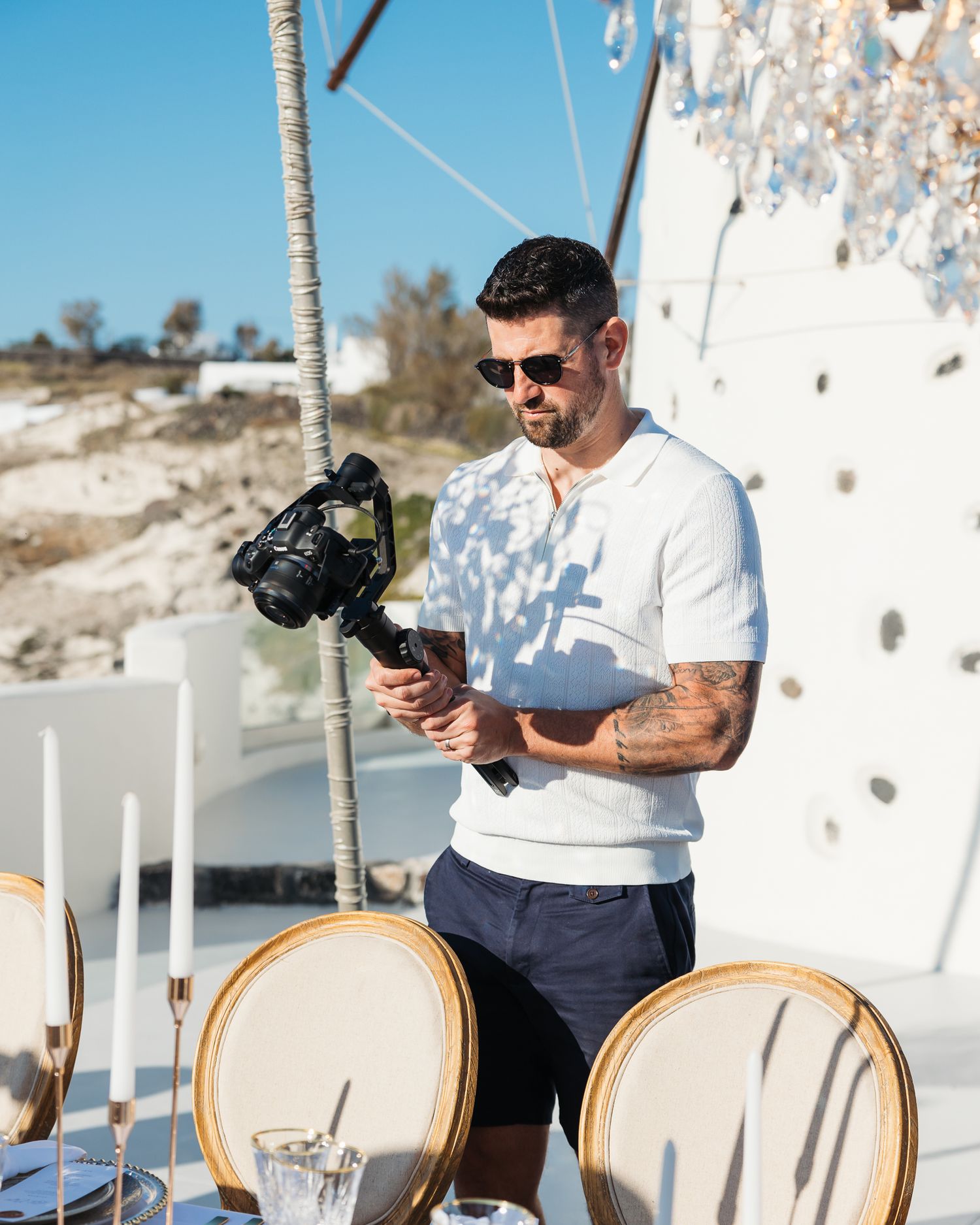 A cameraperson with sunglasses adjusts equipment on sunny outdoor deck with elegant place settings.
