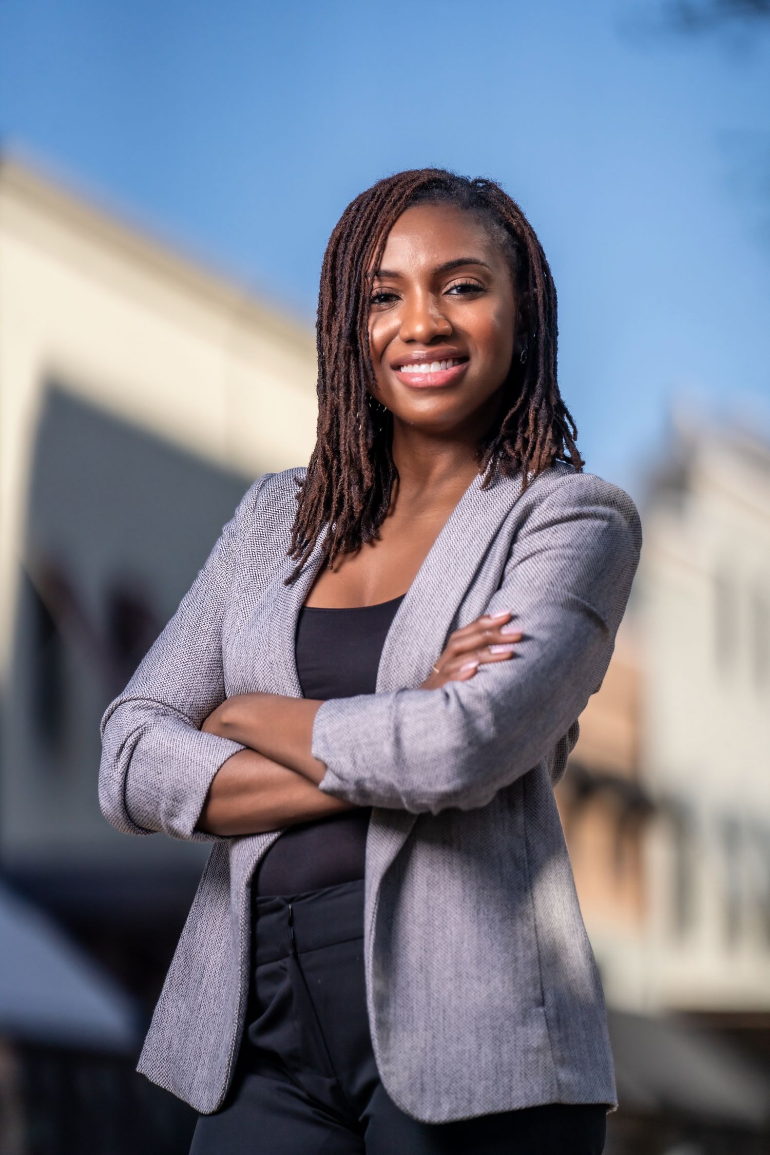 Professional wearing a gray blazer with dark hair smiles confidently with arms crossed against urban background.