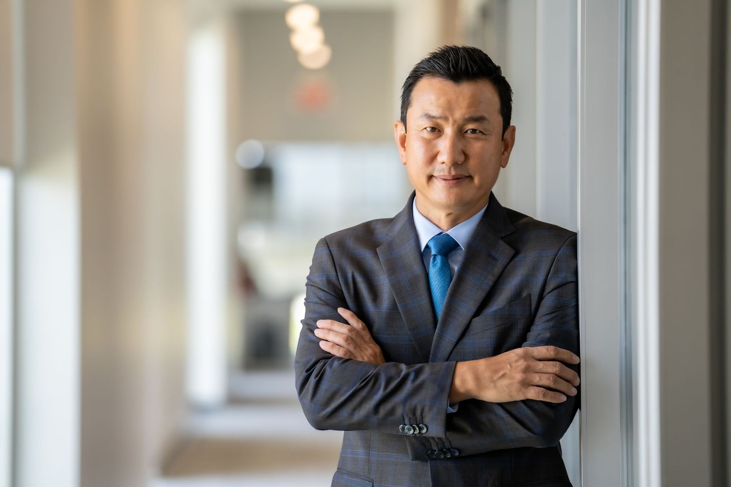 Professional businessman in gray suit and blue tie stands confidently with arms crossed in bright office hallway.