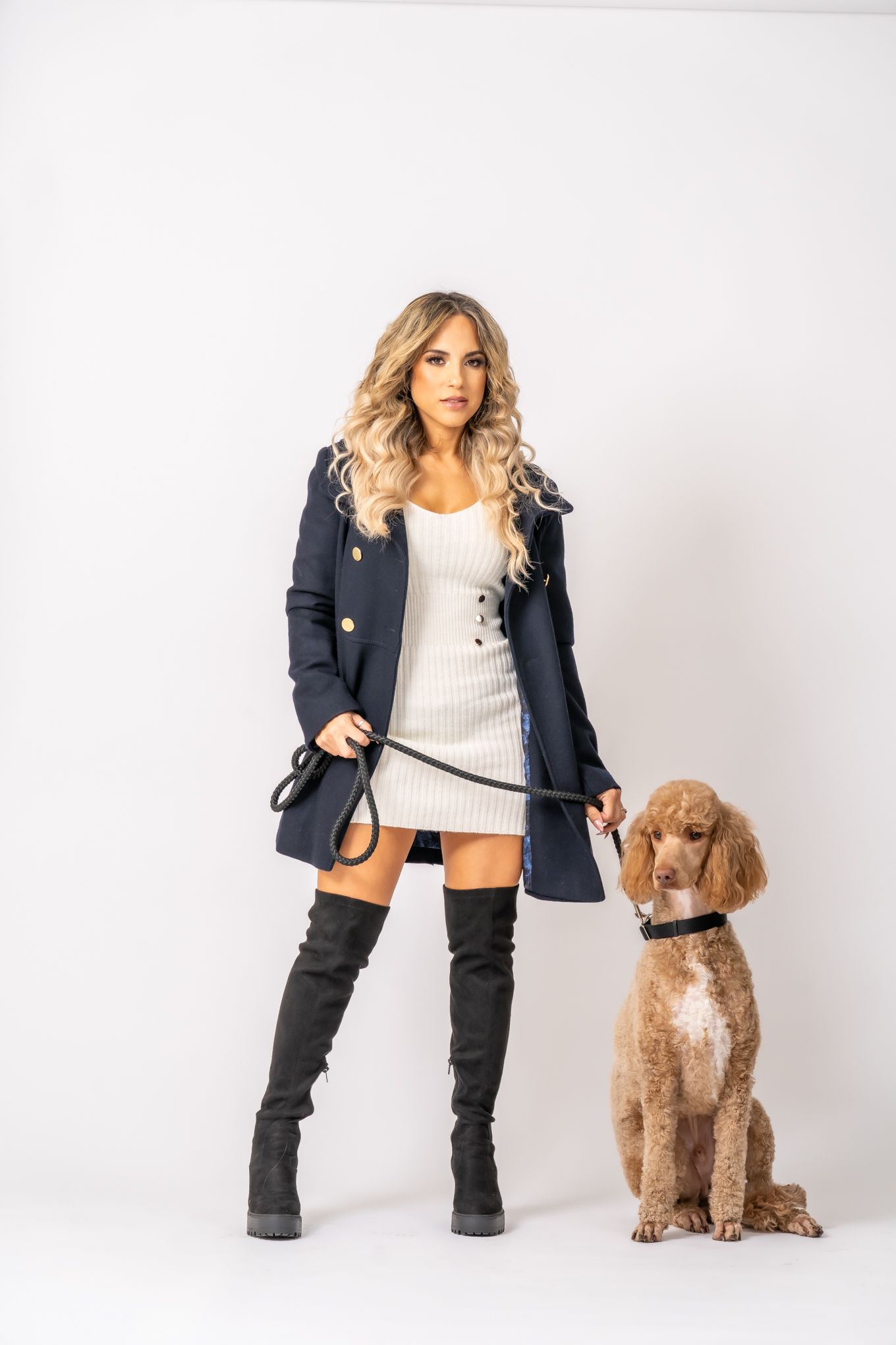 Stylish woman in navy coat and boots poses with well-trained goldendoodle dog against white background.