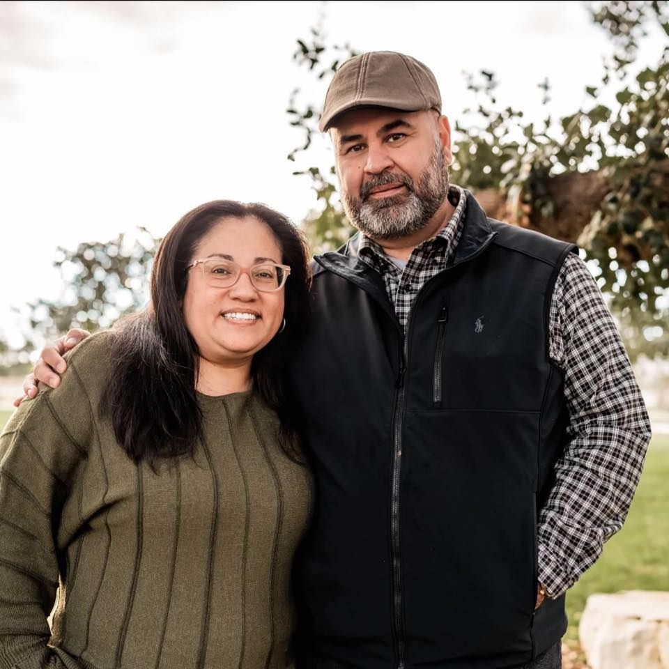 A couple smiling together outdoors, with one wearing a sweater and one in a black vest and checkered shirt.