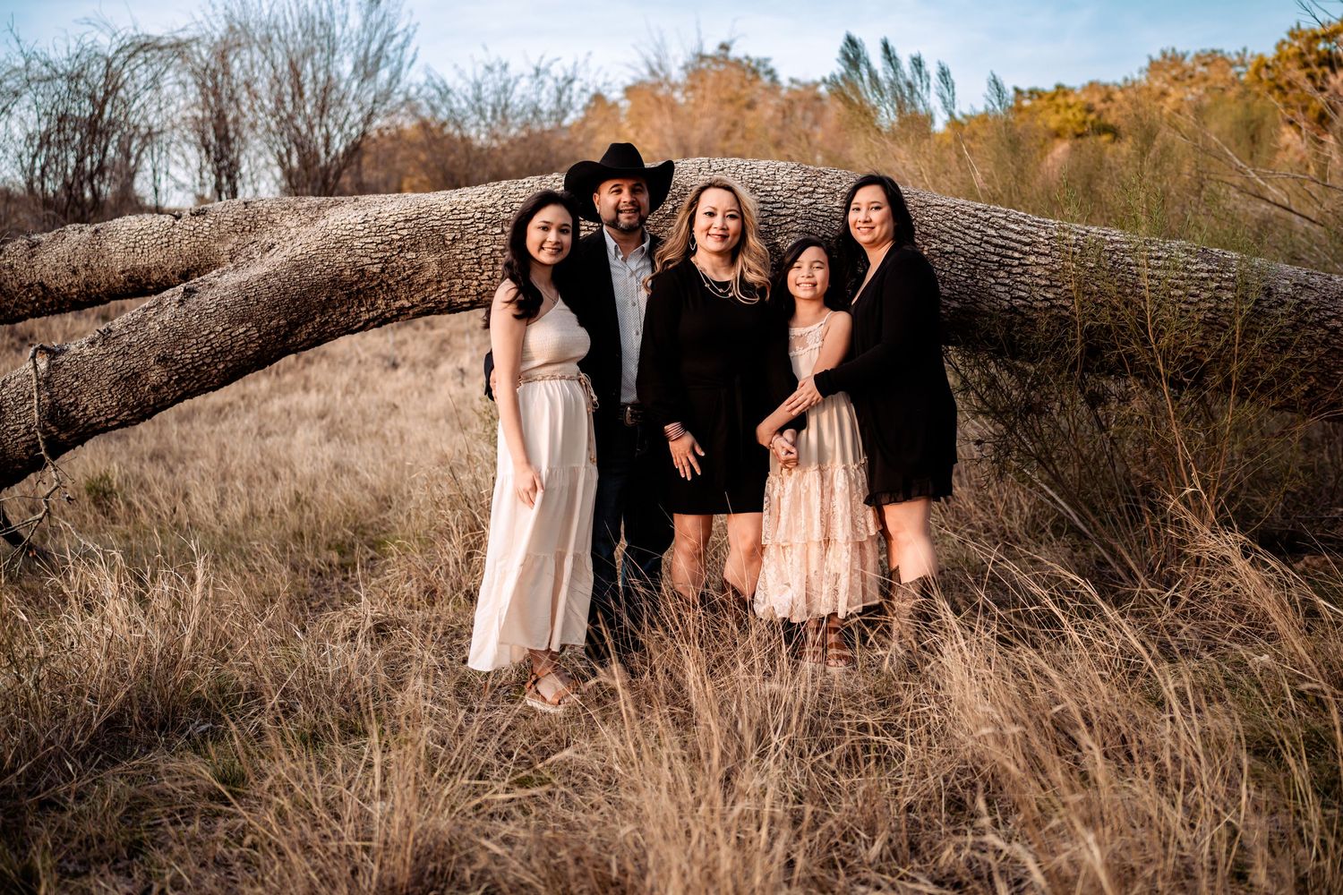 A group of people dressed for a formal occasion pose together on dried grass near a large fallen tree in autumn.