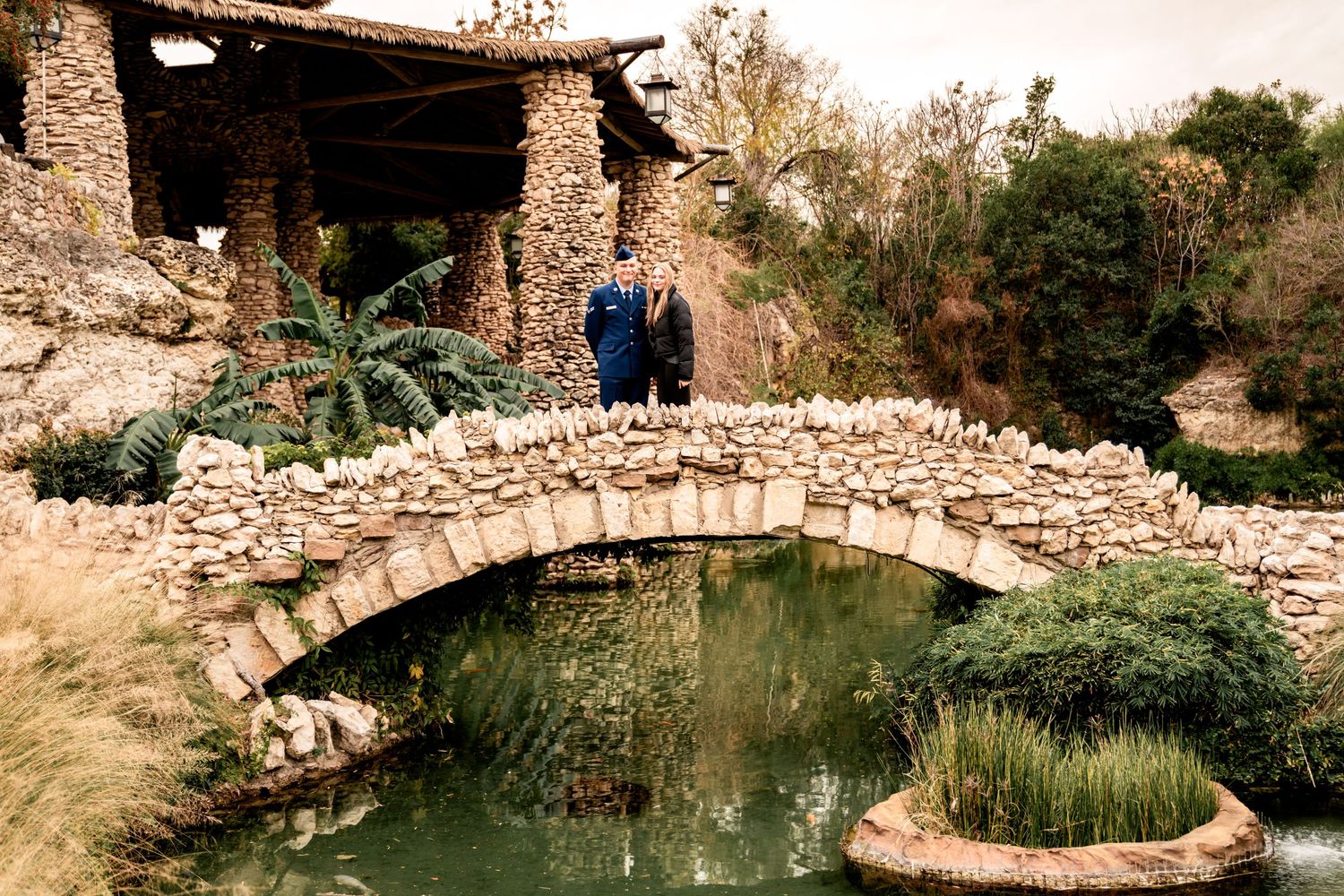 Stone arch bridge spans a tranquil garden pond near a rustic stone building in a natural setting.