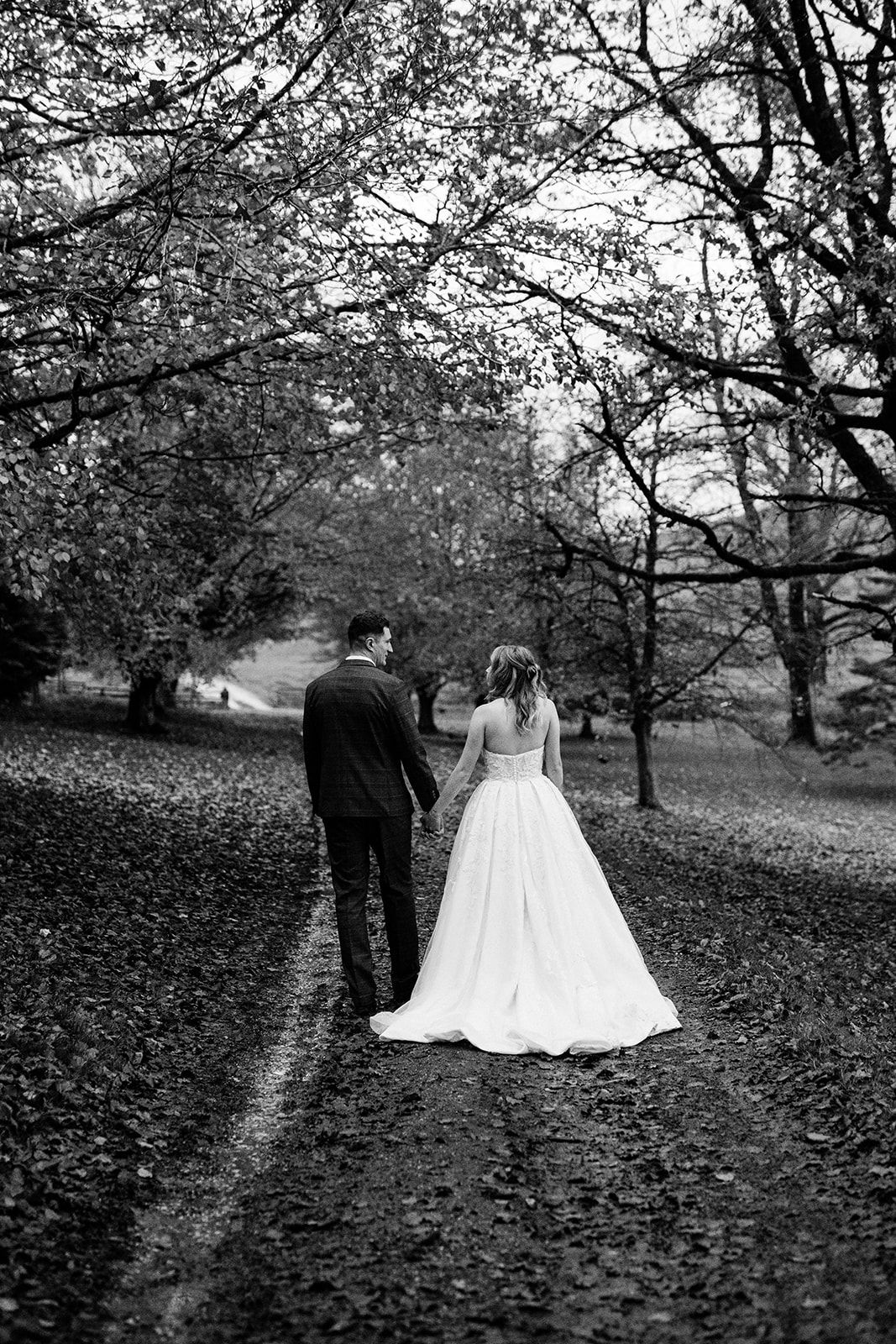 Black and white photo of a bride and groom holding hands as they walk away along a leaf-covered path beneath autumn trees.