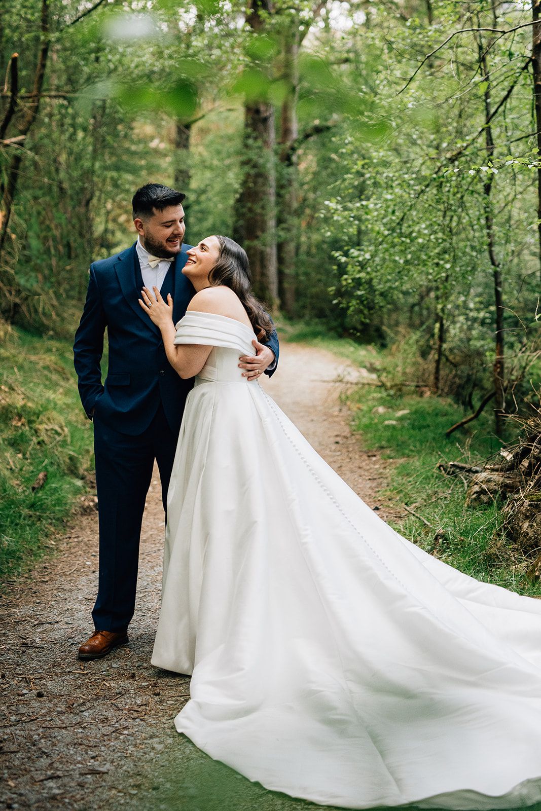 A romantic wedding couple embraces on a wooded path as the bride's elegant white gown flows behind them.