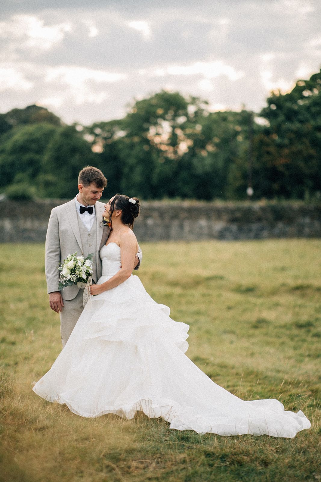 Bride in a flowing white tiered gown and groom in a light gray suit with bow tie embrace in a grassy field at sunset