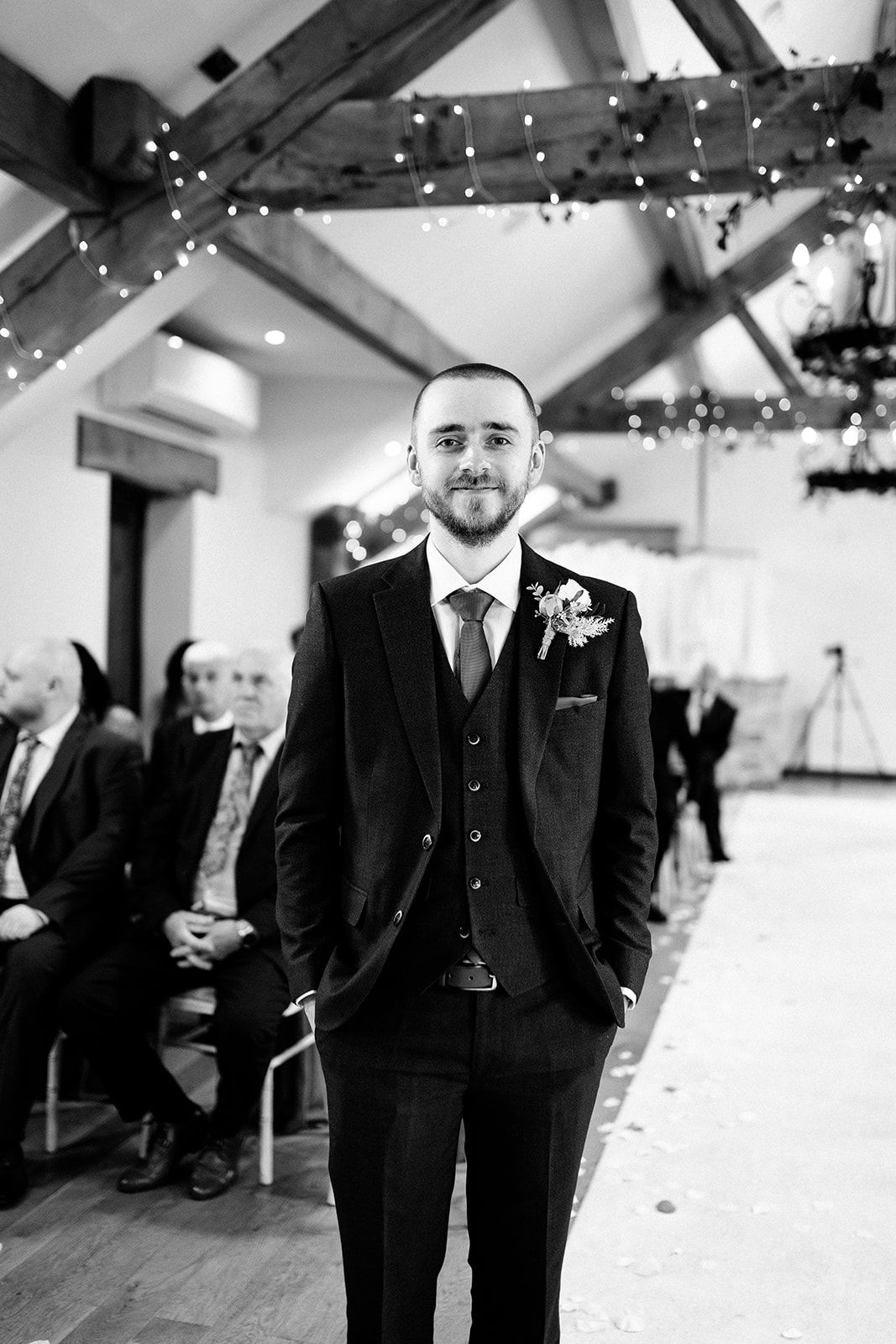Groom in a three-piece tweed suit with floral boutonniere stands at the aisle of a rustic barn wedding venue with string lights.
