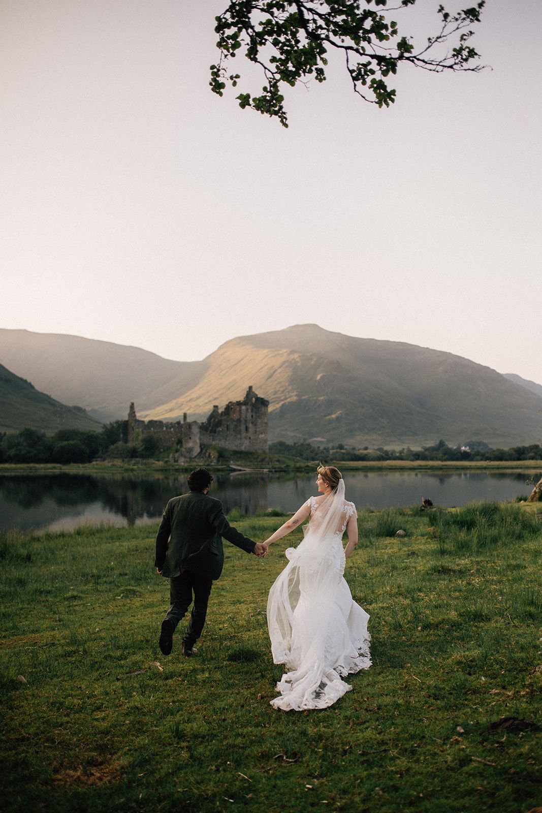 Bride and groom holding hands walking toward a loch and castle ruins with mountain backdrop at sunset.