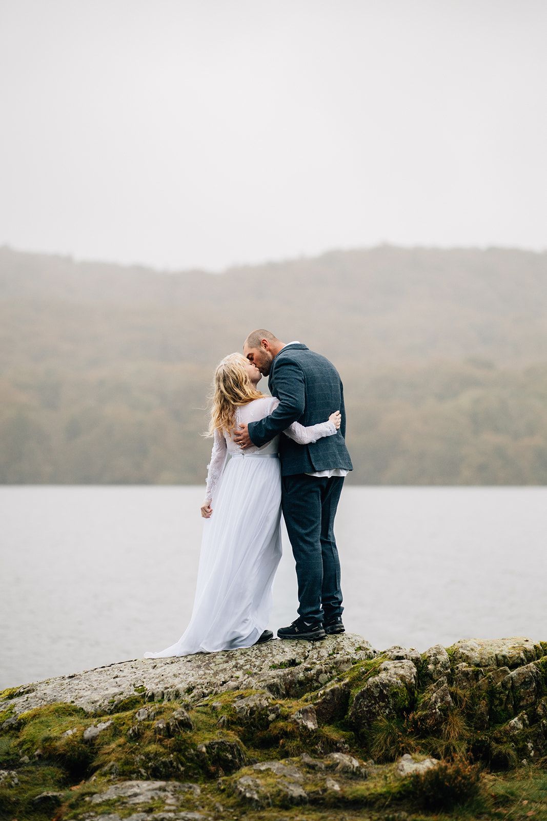A couple shares an intimate moment on a rocky hilltop overlooking a misty lake in the Scottish Highlands.