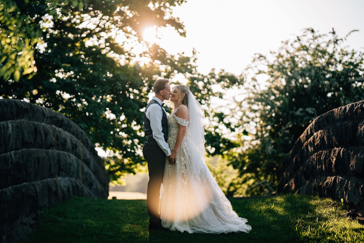 A couple in wedding attire poses together in a golden sunset garden setting with lush greenery and trees.