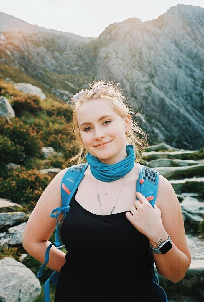 A hiker wearing a blue neck gaiter and backpack poses against a backdrop of rugged mountain scenery.