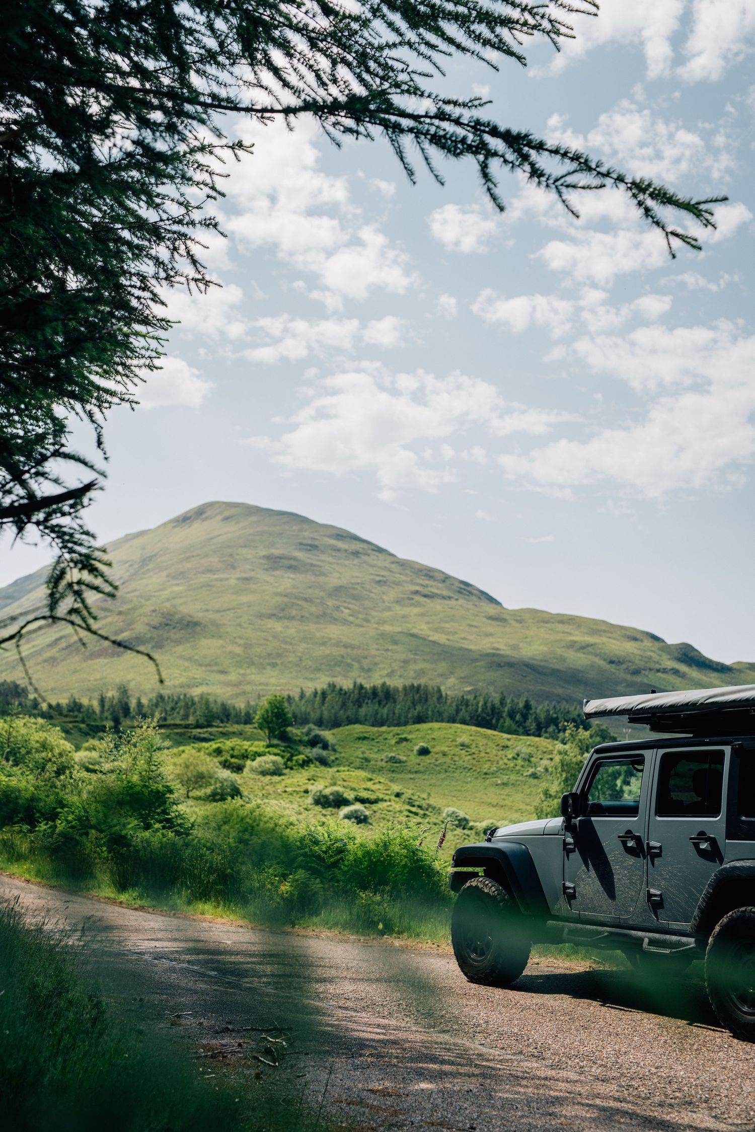 A black Jeep Wrangler parked on a gravel path with rolling green Scottish hills and evergreen trees in the background.