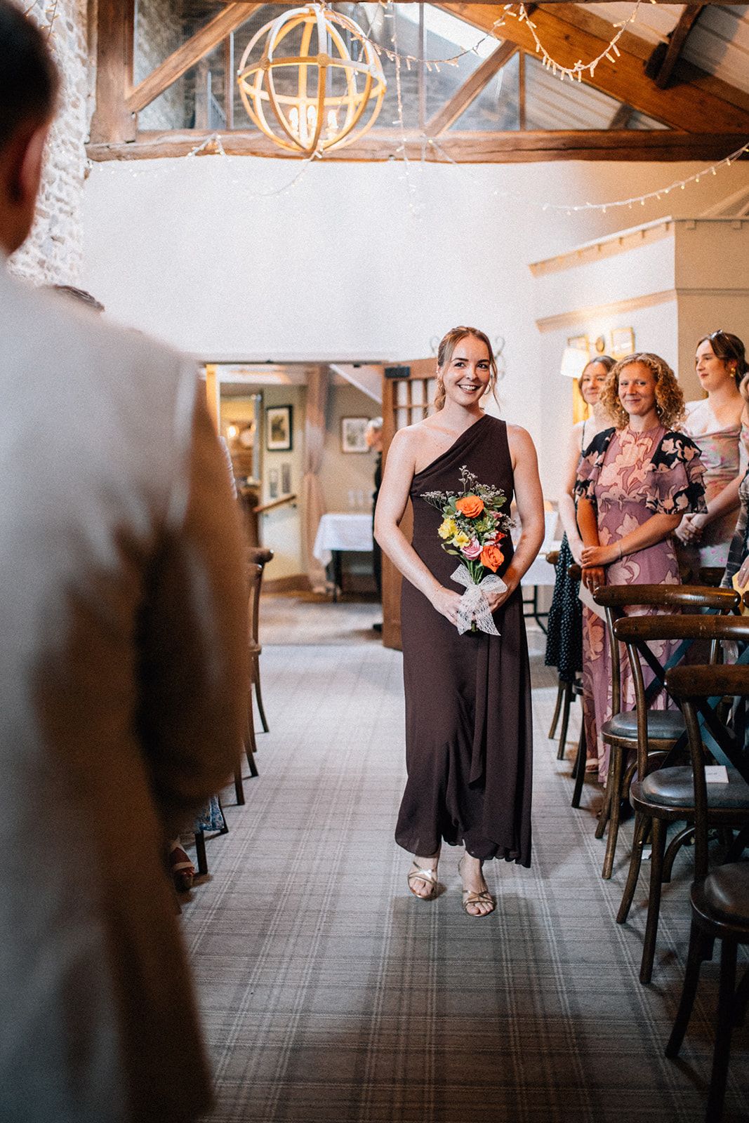 Smiling bridesmaid in a plum dress carries an orange and yellow bouquet down the aisle of a rustic wedding venue with wooden beams.