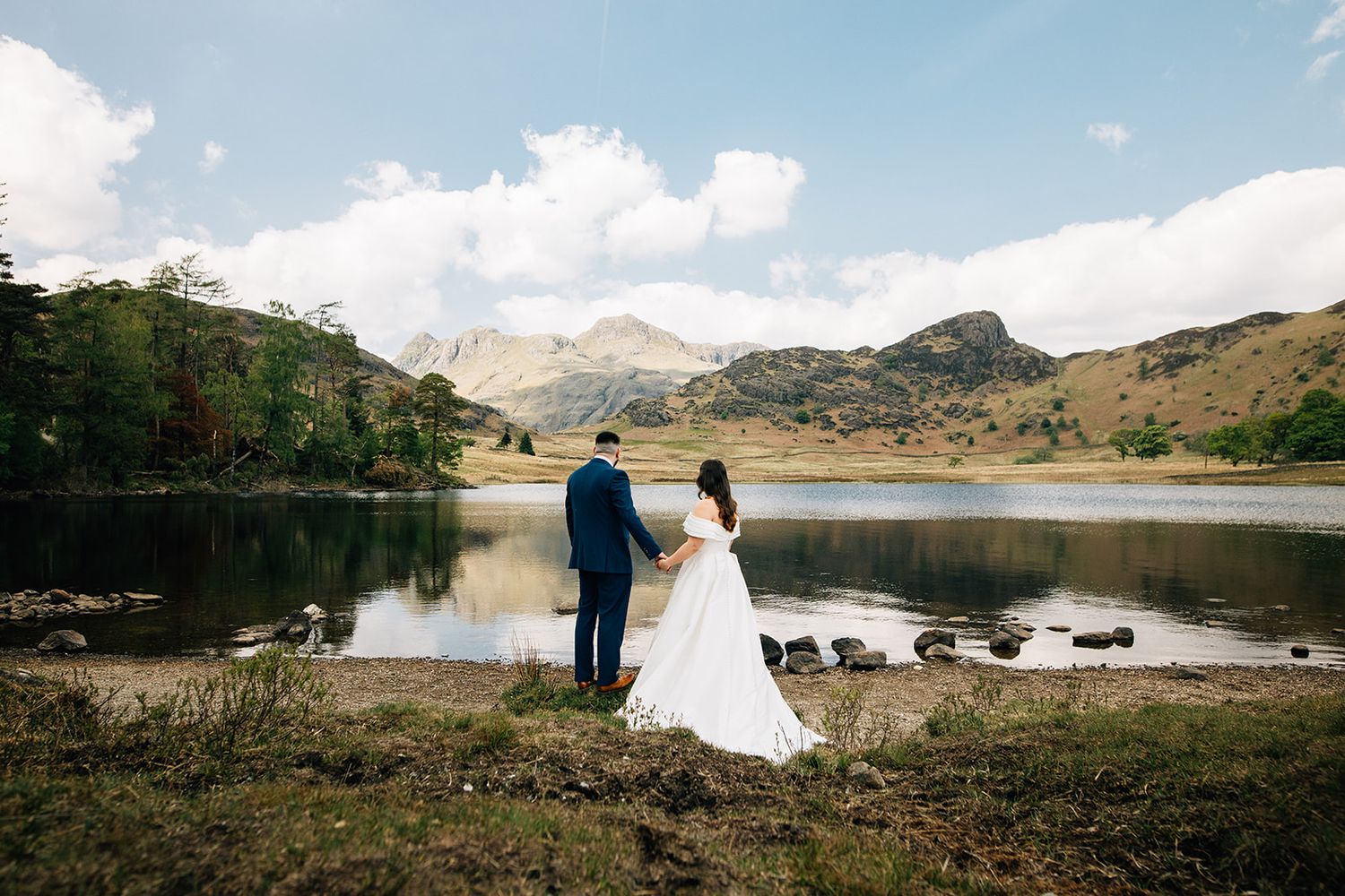 Couple in wedding attire stands by a serene mountain lake surrounded by rugged peaks and natural scenery.
